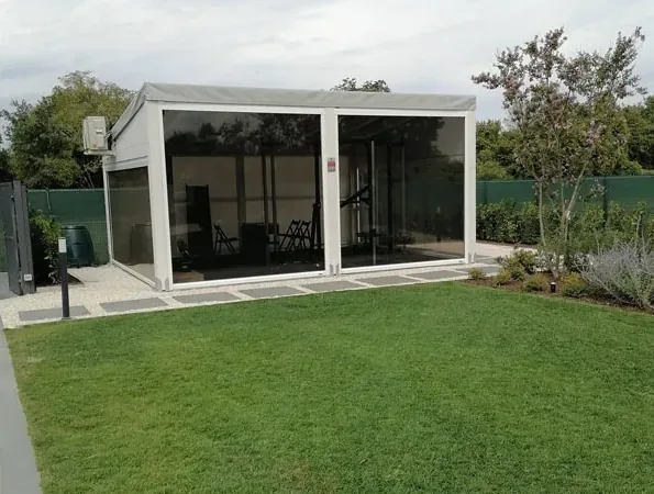 Dopo: pergola bianca chiusa da vetrate davanti al prato, con un alberello e arbusti accanto