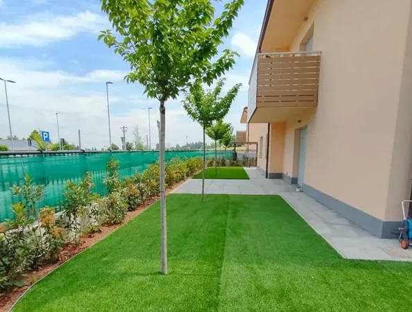 Dopo: prato sintetico con una fila di alberelli giovani e una siepe giovane lungo la recinzione, accanto alla casa