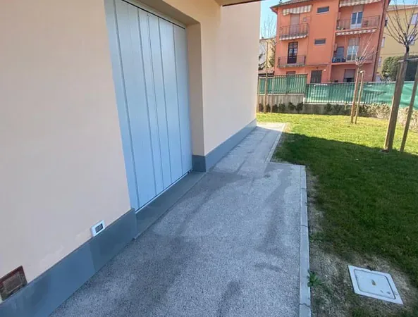 Prima: vialetto in cemento lungo il garage, con il prato sulla destra e un pozzetto a terra
