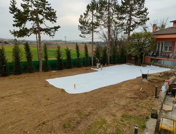 Prima: terreno spoglio con un telo bianco steso e la fila di conifere a cono lungo la recinzione, sotto alcuni alberi alti