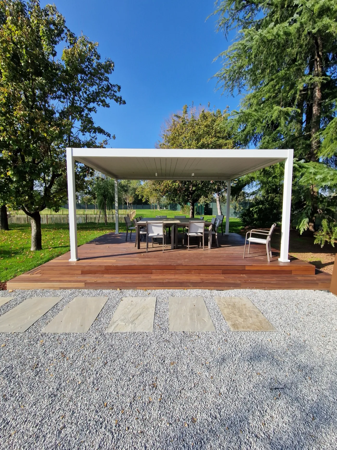 Pergola bianca su una pedana in legno in fondo a un giardino, con un camminamento di lastre sulla ghiaia
