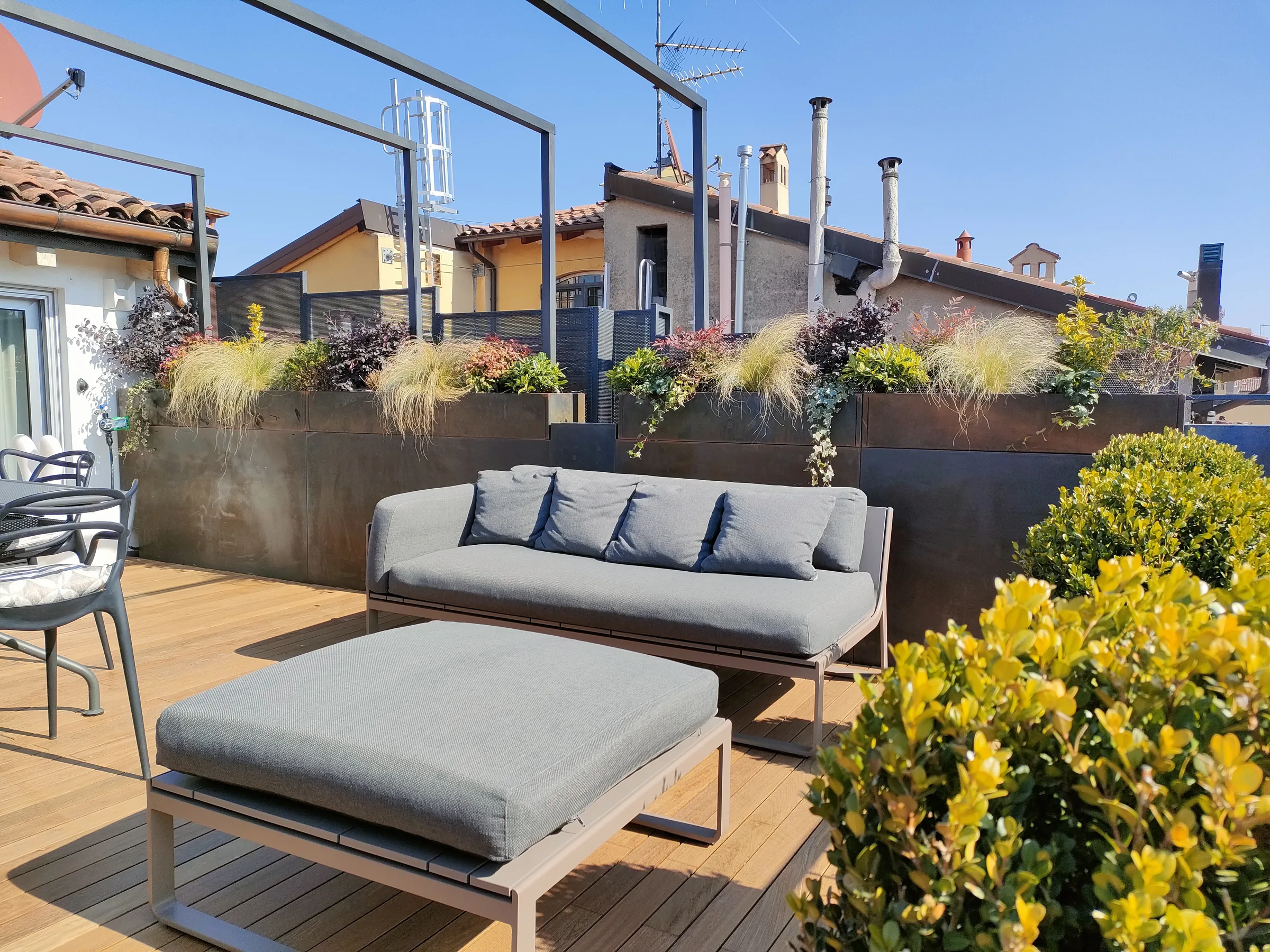 Terrazzo con fioriere in corten piene di graminacee e arbusti, e un divano grigio sulla pedana in legno