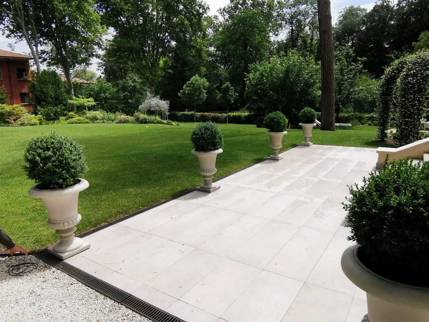 Vialetto in lastre chiare fra il prato e i vasi con sfere di bosso, verso il giardino alberato