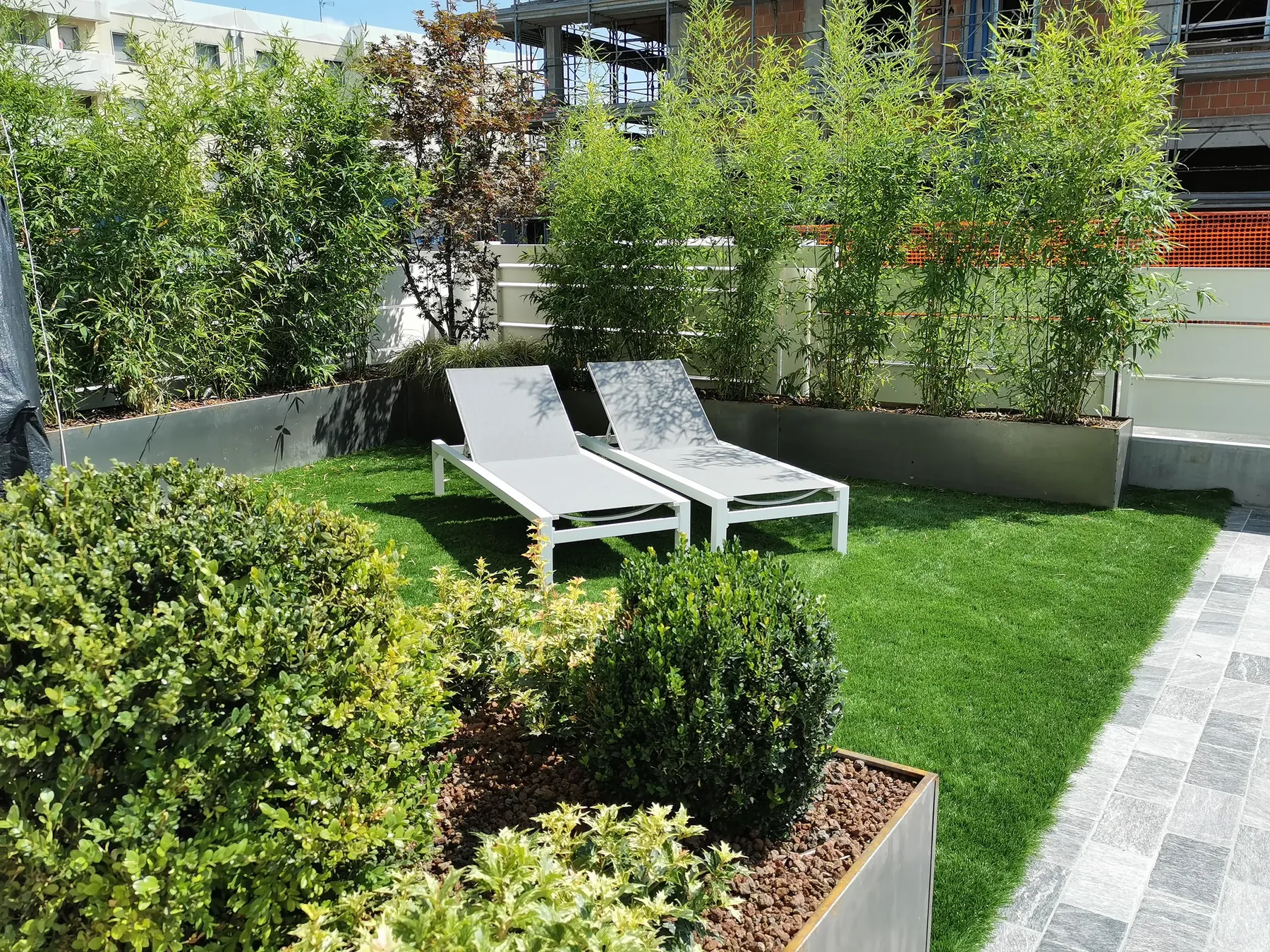 Giardino con prato sintetico e due lettini bianchi, chiuso da fioriere grigie piantate a bambù, con cespugli di bosso in primo piano