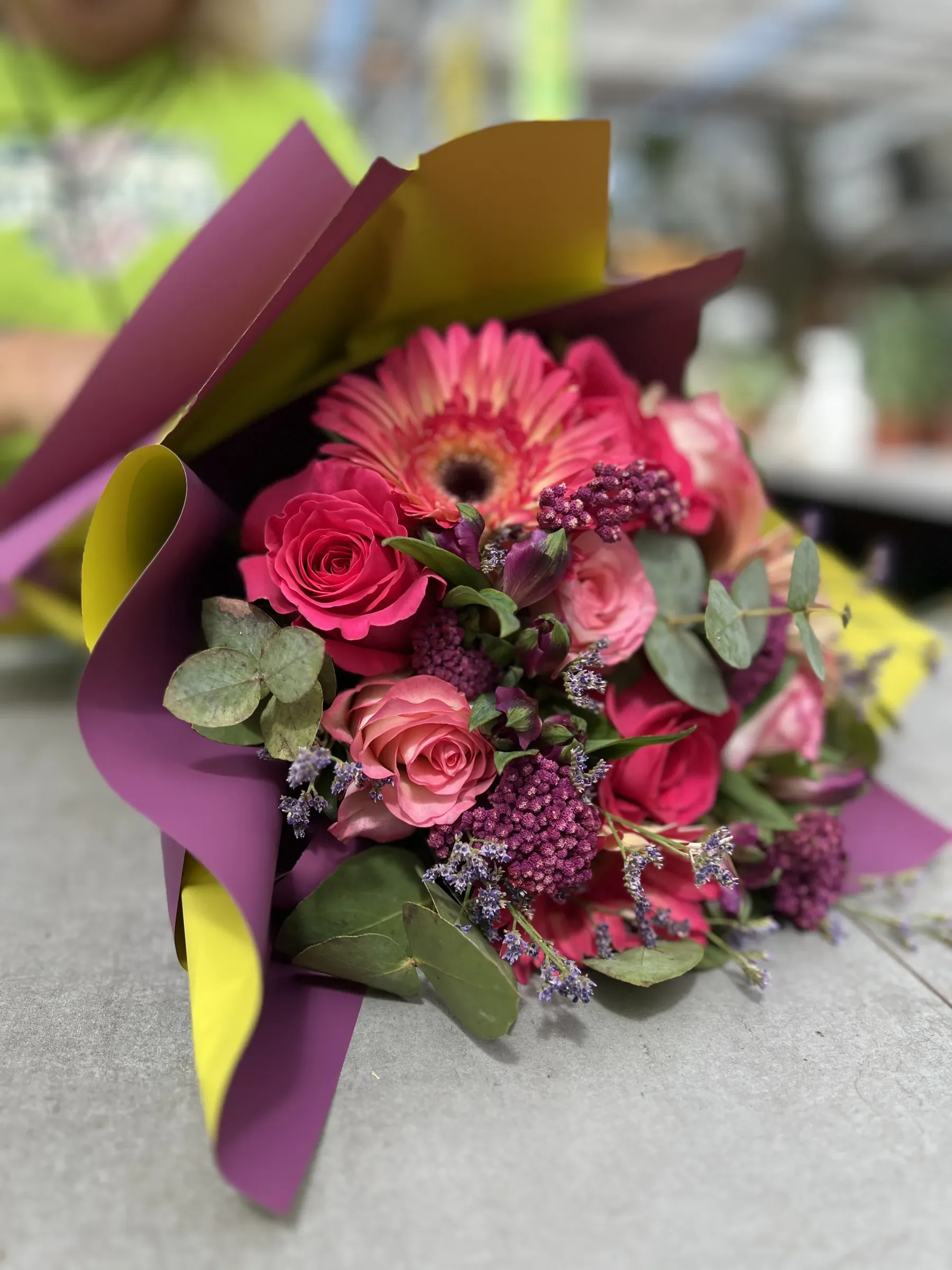 Mazzo con una gerbera, rose rosa e fucsia, limonium viola ed eucalipto, incartato in carta viola e gialla