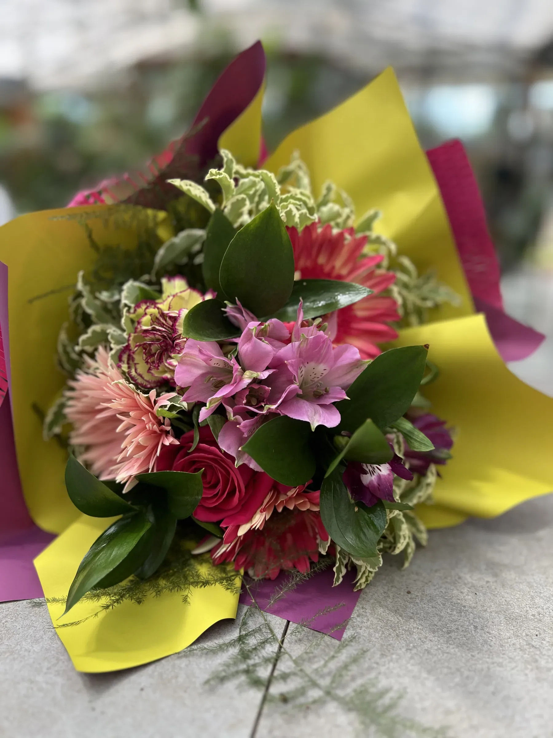 Mazzo di alstroemerie rosa, gerbere, una rosa fucsia e un garofano screziato, con foglie variegate, in carta gialla e fucsia, appoggiato sul banco