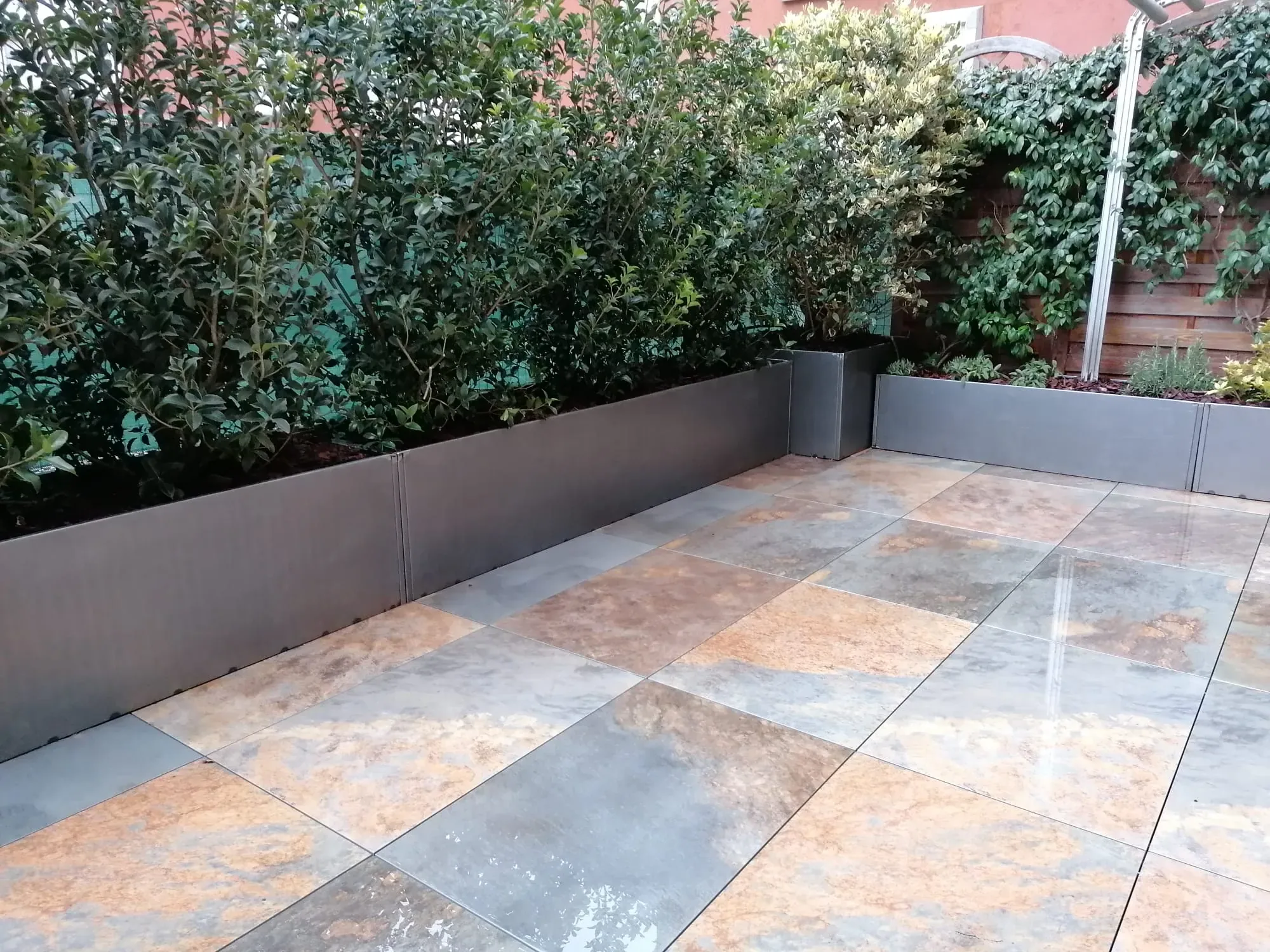 Terrazzo pavimentato con grandi lastre dalle sfumature ruggine e grigie, lucide e bagnate, con fioriere metalliche piene di arbusti lungo il bordo