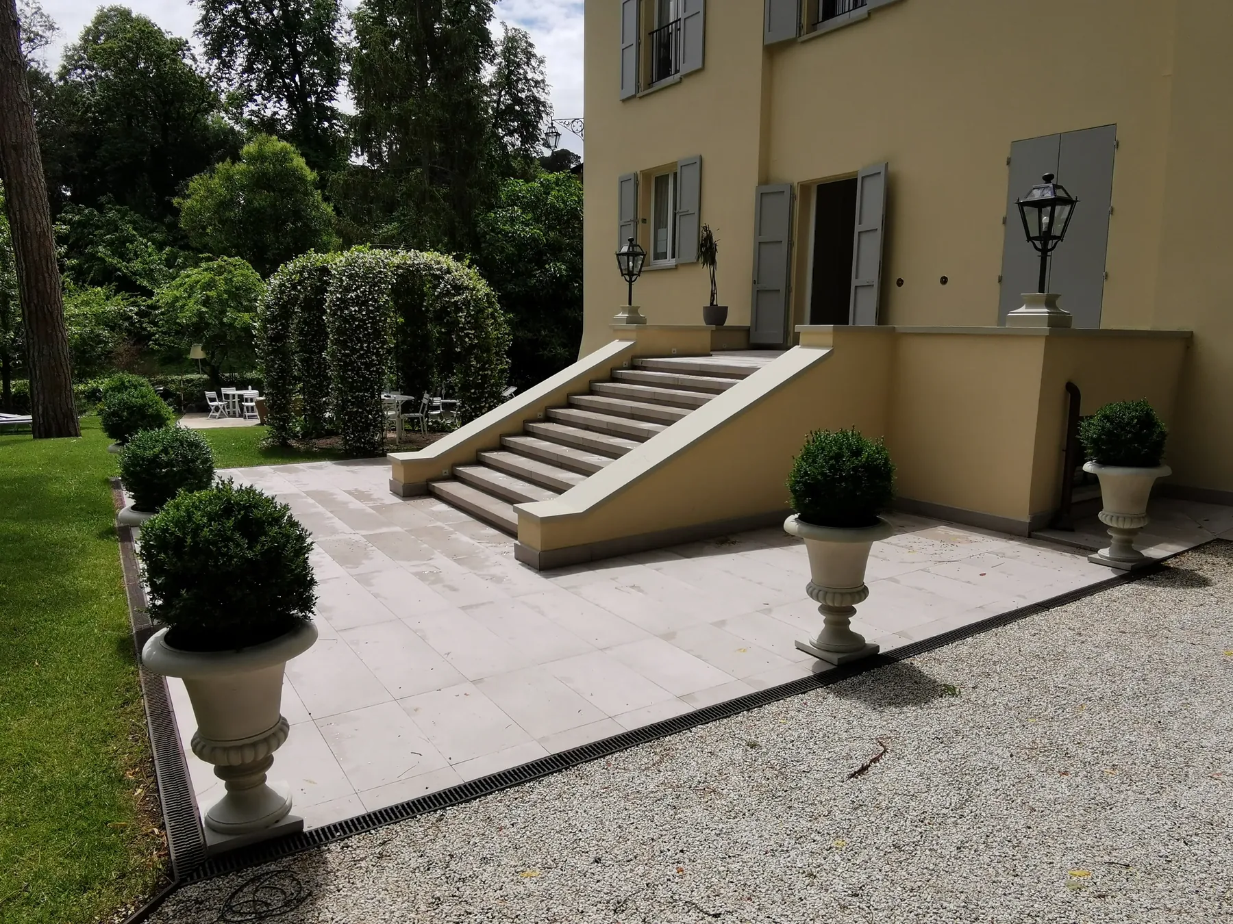 Scalinata di una villa su una terrazza in pietra chiara, con vasi di bosso potati a sfera e archi di rampicanti sul fondo