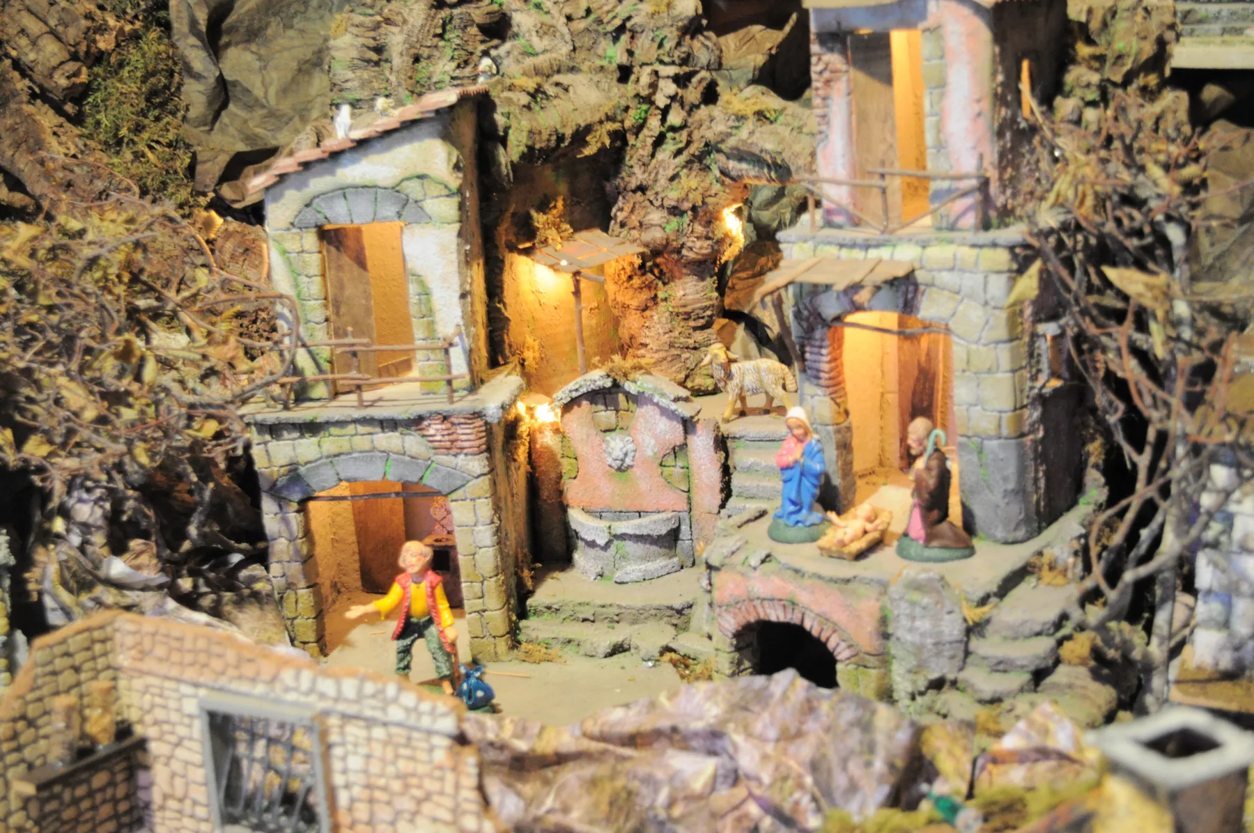 Presepe con case di pietra illuminate, la natività davanti all'ingresso di una casa, una pecora e un pastore sulla strada