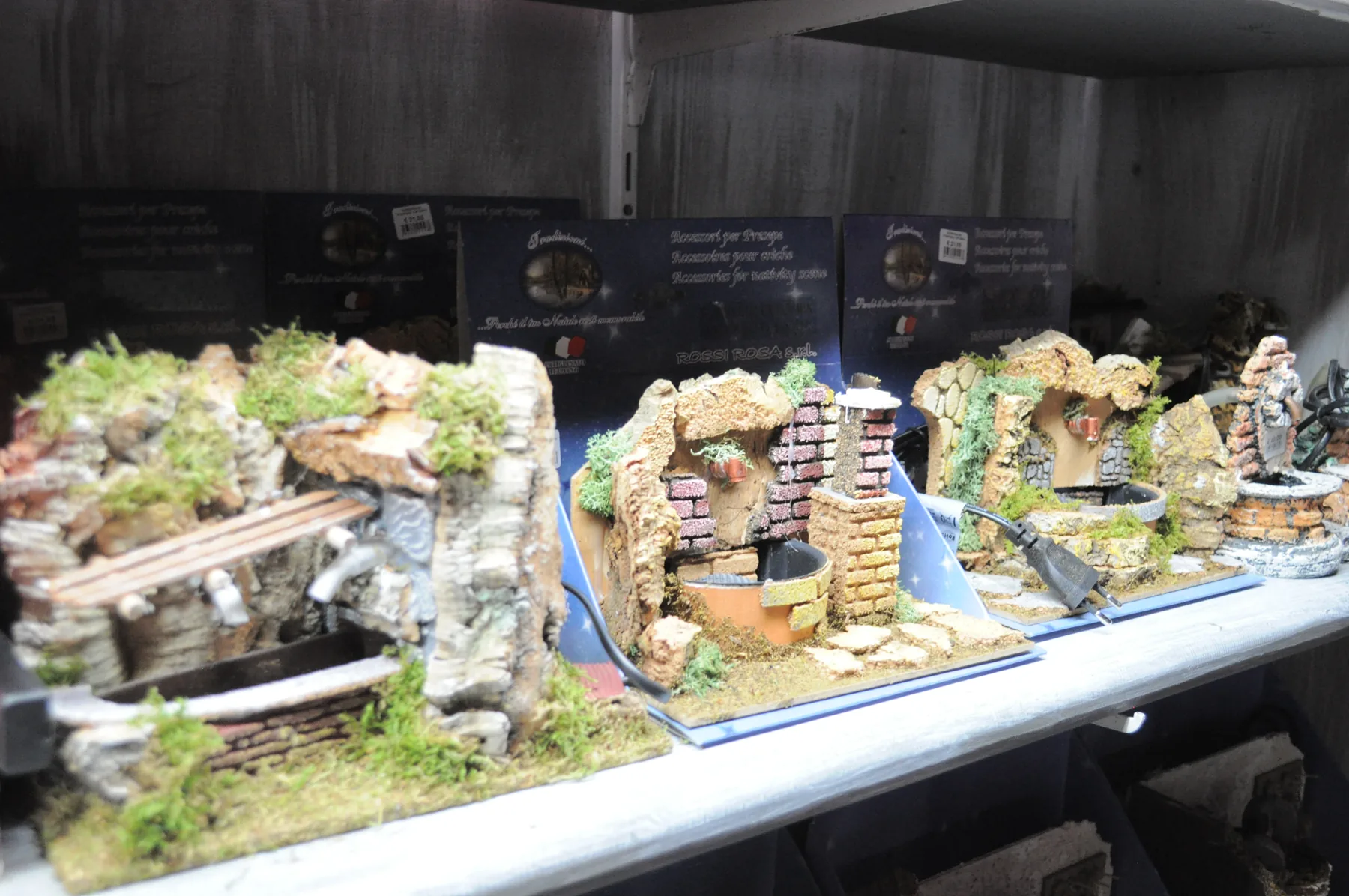 Fontanelle per il presepe fatte di sughero, muschio e muretti di mattoni, allineate su uno scaffale davanti alle scatole degli accessori per presepe
