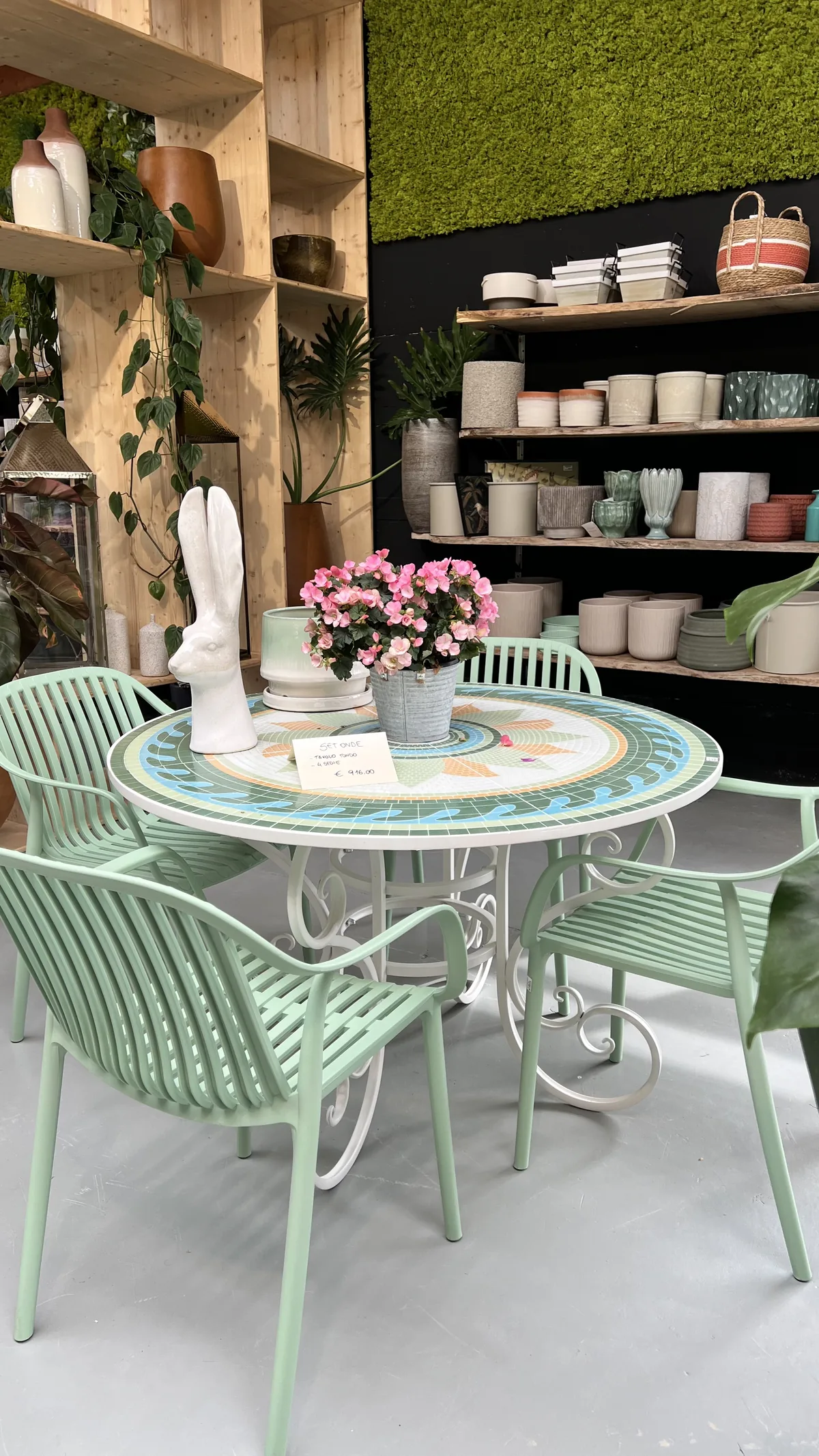 Un tavolo rotondo in mosaico con quattro sedie verde menta davanti agli scaffali dei vasi, con un coniglio bianco decorativo e un vaso di fiori rosa