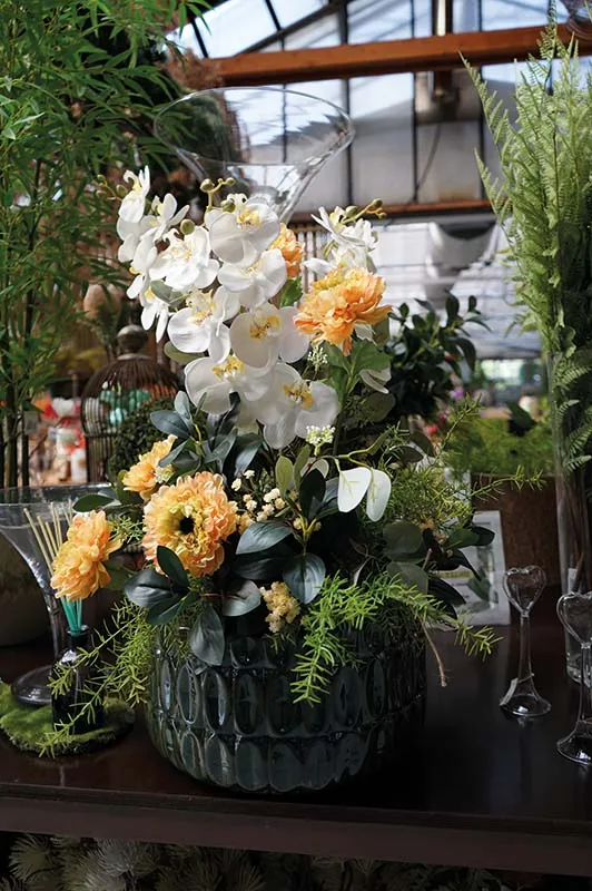 Composizione artificiale di orchidee bianche, fiori arancioni dai petali fitti e felci in un vaso di vetro scuro
