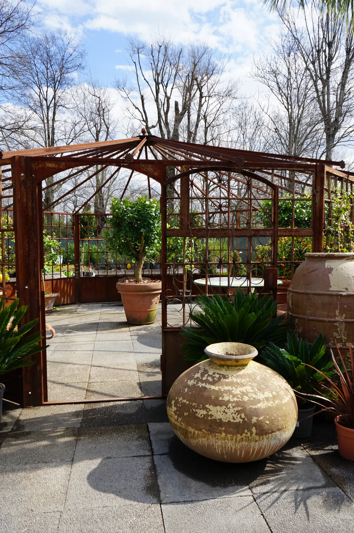 Un gazebo in ferro all'aperto, con un grande orcio di terracotta e piante in vaso sul pavimento in lastre