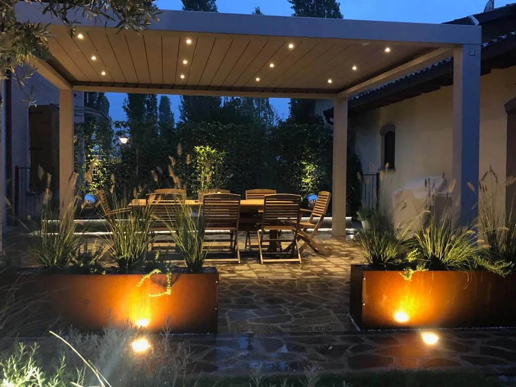 Pergola chiara di sera con i faretti nel soffitto, un tavolo di legno con le sedie e le fioriere in corten illuminate sul pavimento in pietra