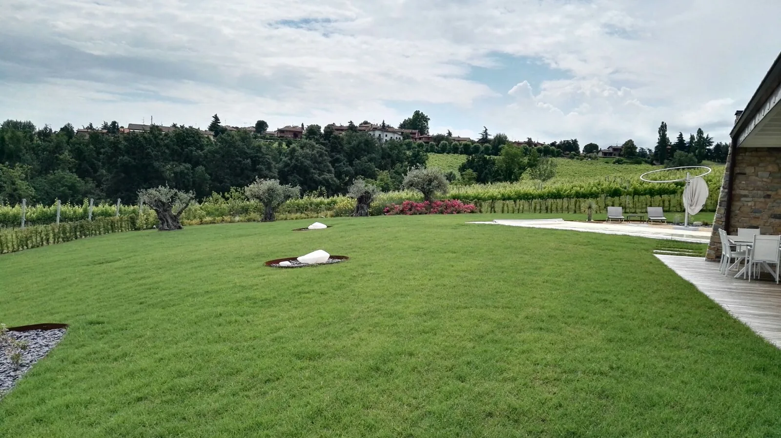 Grande prato in collina con due massi bianchi in aiuole tonde, una fila di olivi sul fondo davanti alle vigne e una pedana in legno con sedie a destra