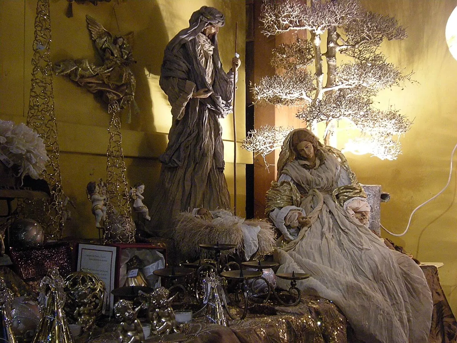 Presepe con le statue di Maria e Giuseppe vestite di tessuti chiari, fra un albero bianco e angeli dorati