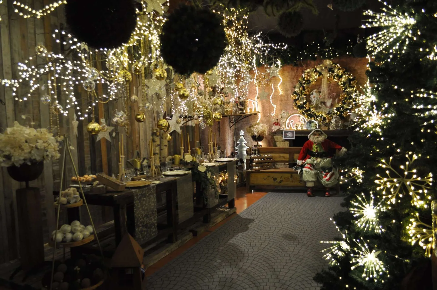 Sala del Villaggio con luci a stella, sfere scure e palline dorate appese sopra una lunga tavola apparecchiata, un Babbo Natale seduto sul fondo e un albero di Natale illuminato in primo piano