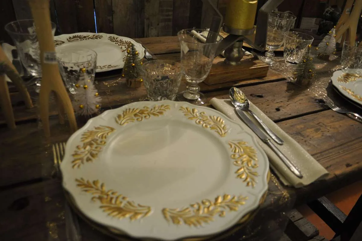 Tavola apparecchiata con piatti bianchi decorati d'oro, bicchieri di cristallo e piccoli alberi innevati