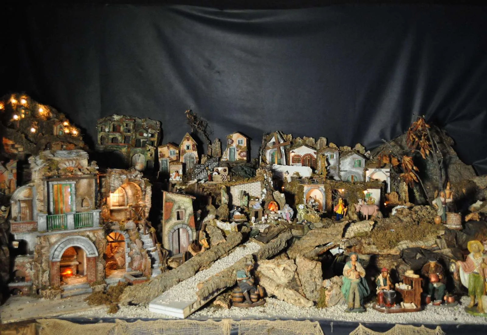 Presepe con case di roccia illuminate, tronchi, sentieri e statuine di pastori, su un fondale scuro