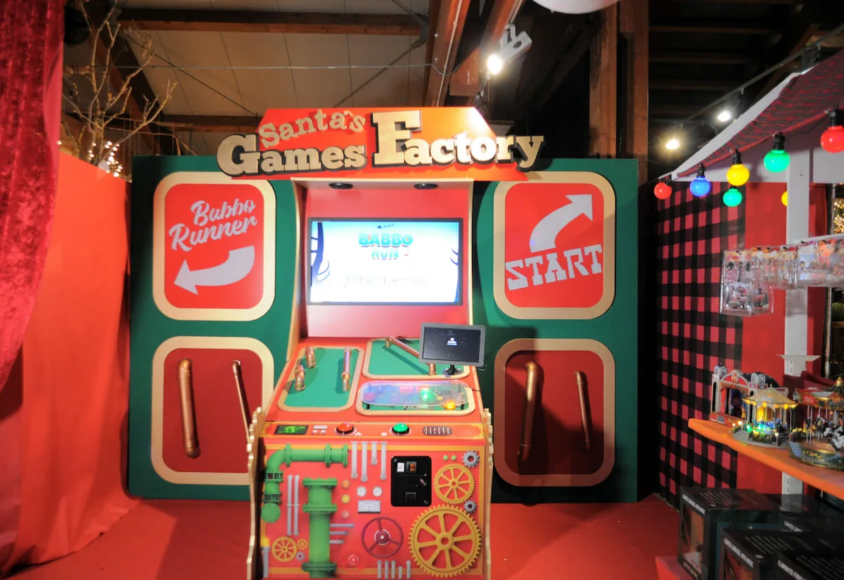 Il gioco a schermo della Santa's Games Factory, con la console rossa a leve e ingranaggi e le frecce Babbo Runner e Start