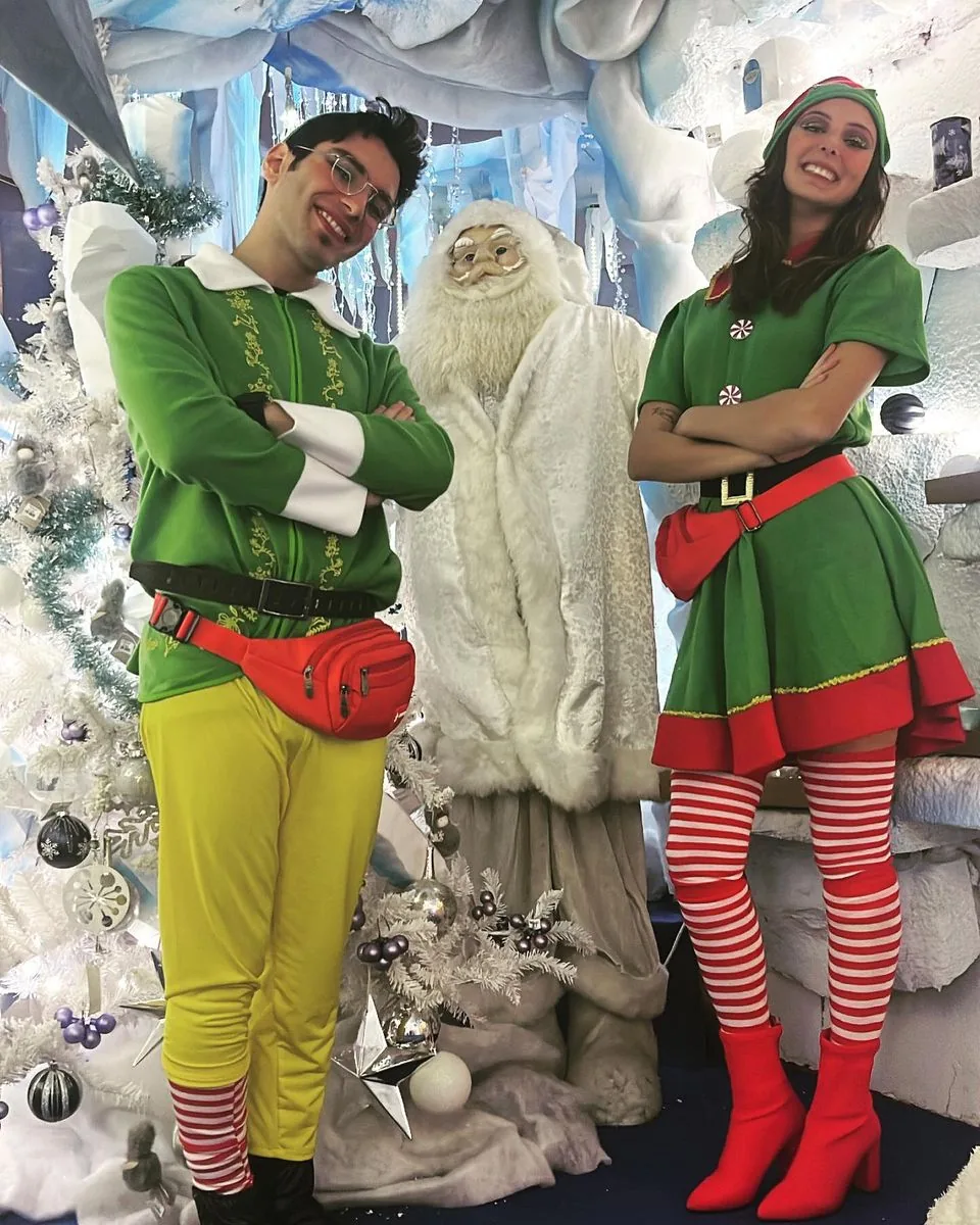 Due animatori vestiti da elfi a braccia conserte accanto a una statua di Babbo Natale bianco