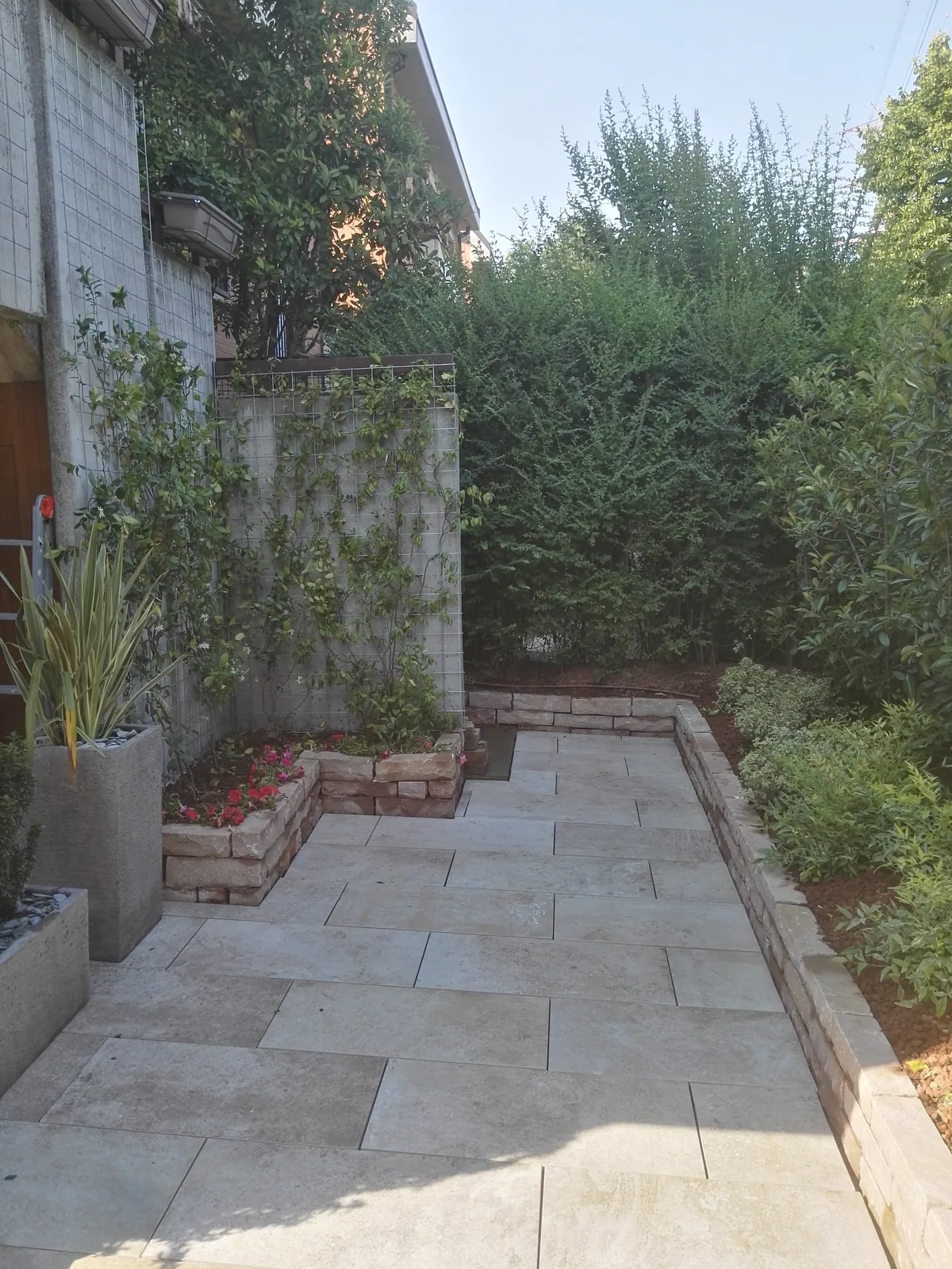 Vialetto in lastre di pietra chiara fra due bordure in blocchetti di pietra, con piante in vaso e una siepe alta
