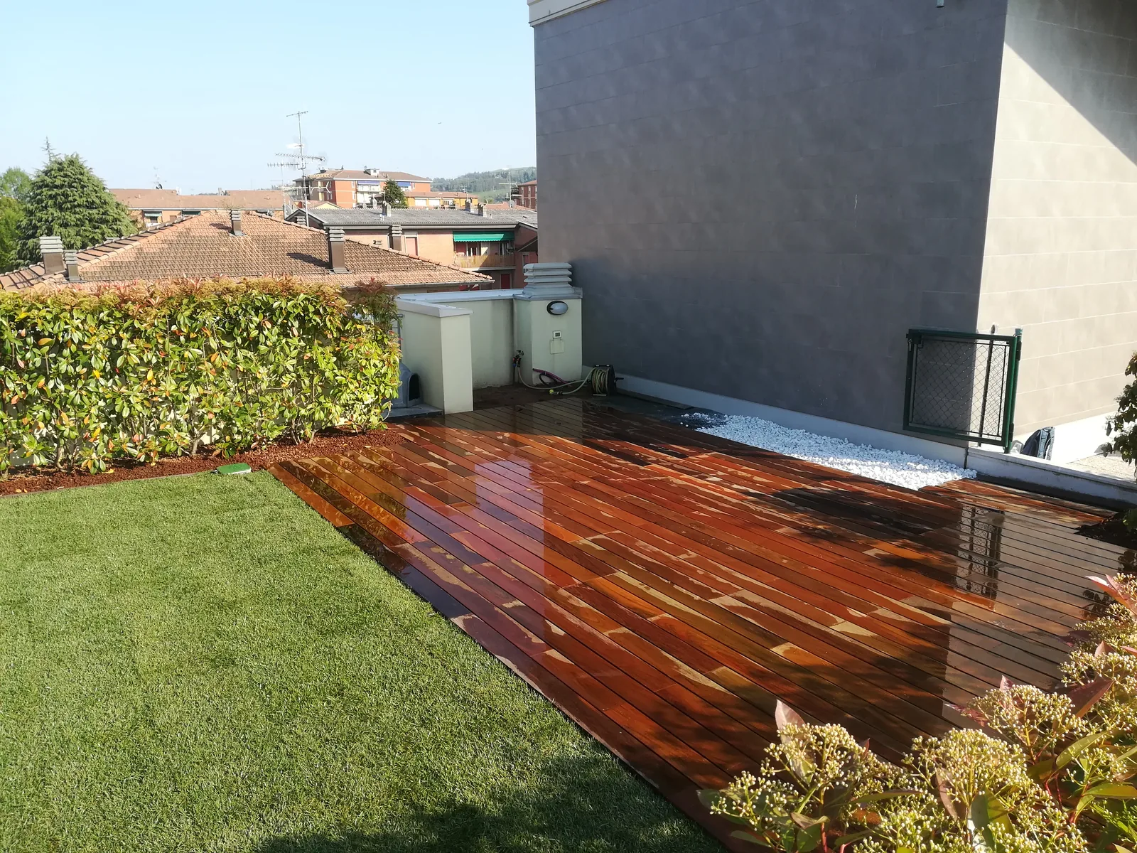 Pedana in legno affiancata a un prato appena posato, su un terrazzo con una siepe dalle punte rosse sul bordo e una striscia di ciottoli bianchi