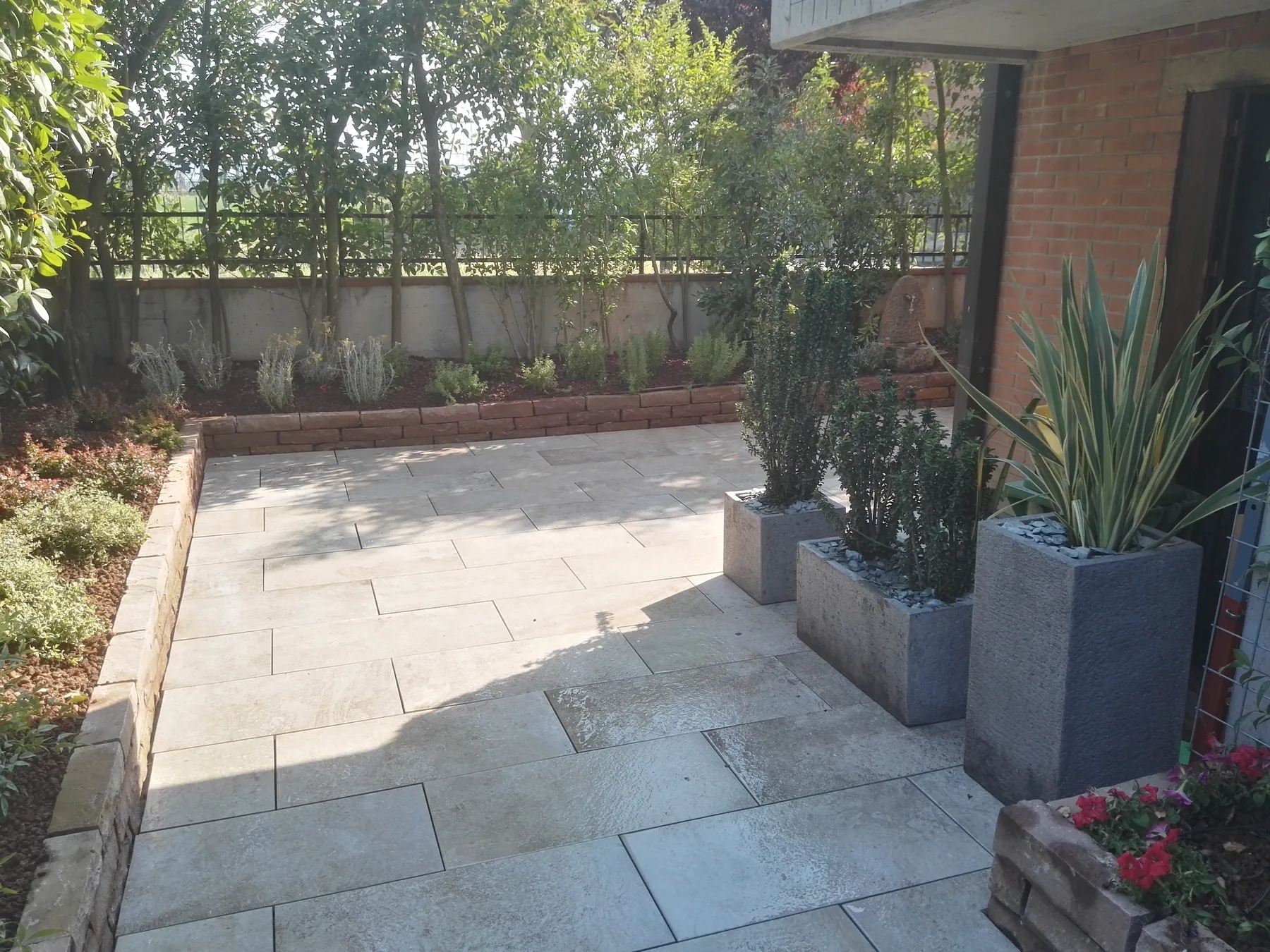 Cortile pavimentato in grandi lastre di pietra chiara, con vasi alti grigi di arbusti sempreverdi e phormium e un'aiuola bordata da un muretto di blocchi lungo il muro