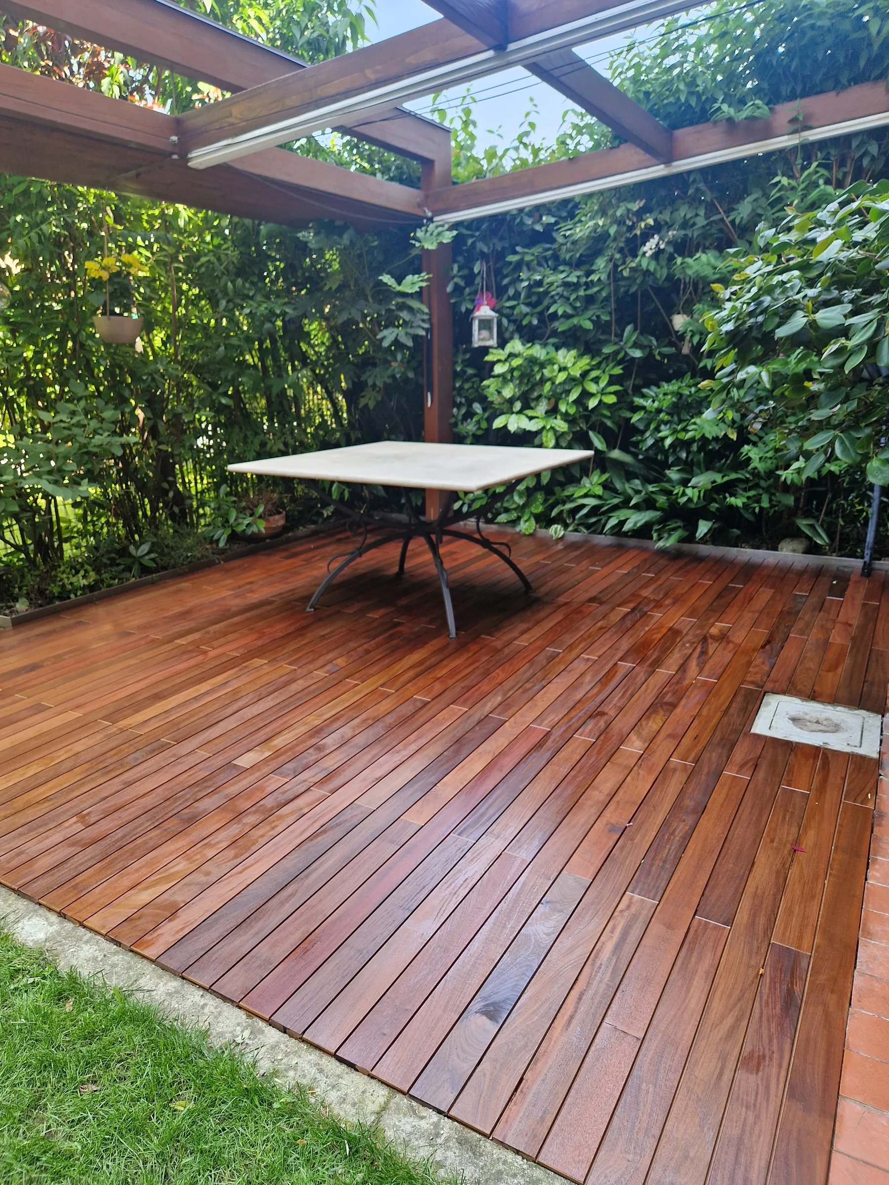Pedana in listoni di legno sotto una pergola di legno circondata dal verde, con un tavolo dal piano chiaro e il prato sul bordo