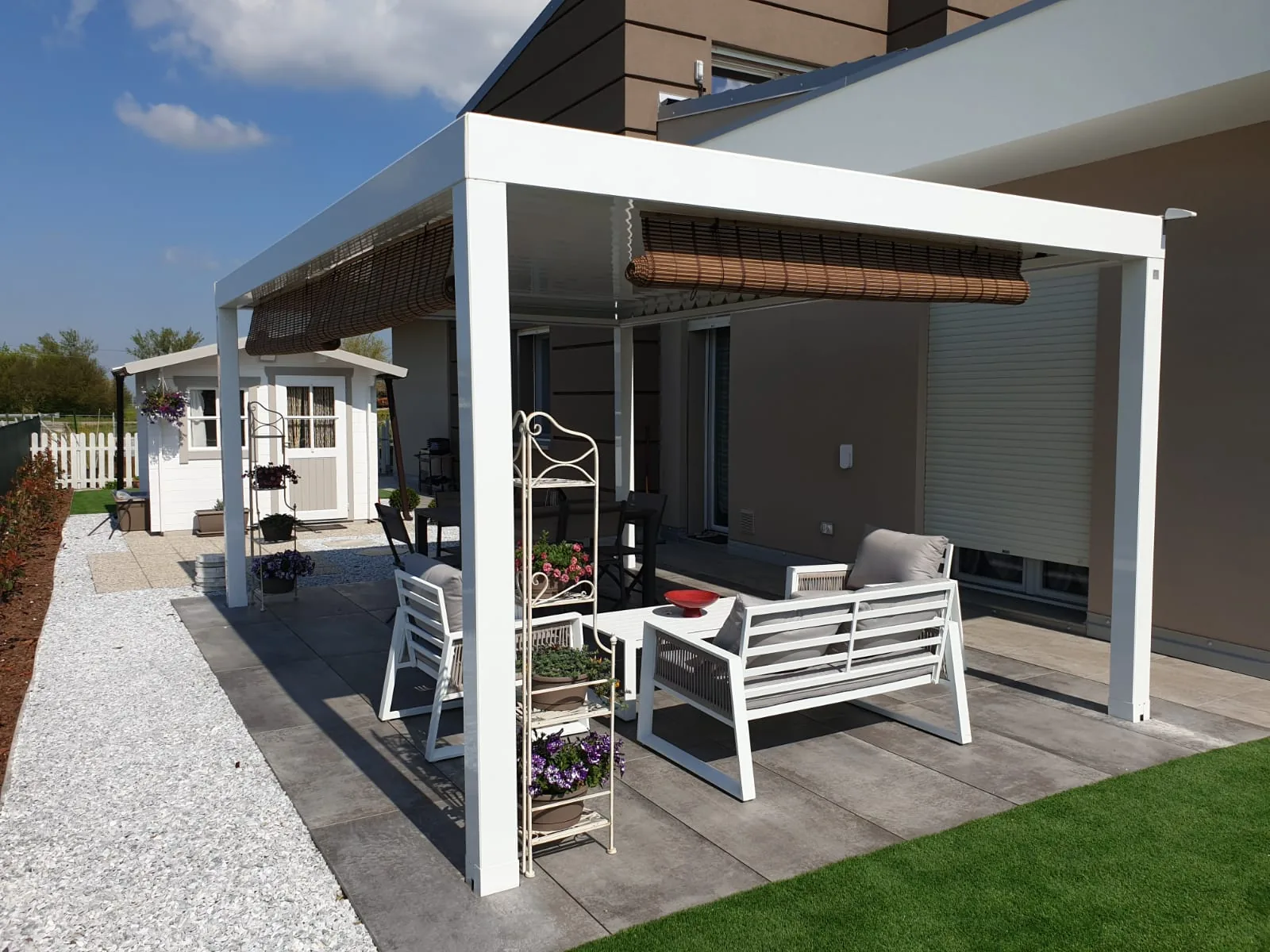 Pergola bianca con le tende a rullo sopra un salotto da esterno, su un pavimento scuro fra il vialetto in ghiaia e il prato