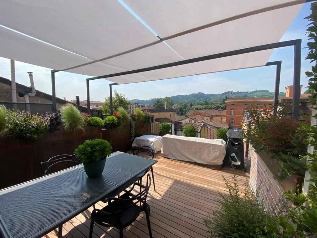 Terrazzo con una tenda bianca tesa su un telaio scuro, tavolo lungo, pavimento in legno e fioriere lungo il parapetto