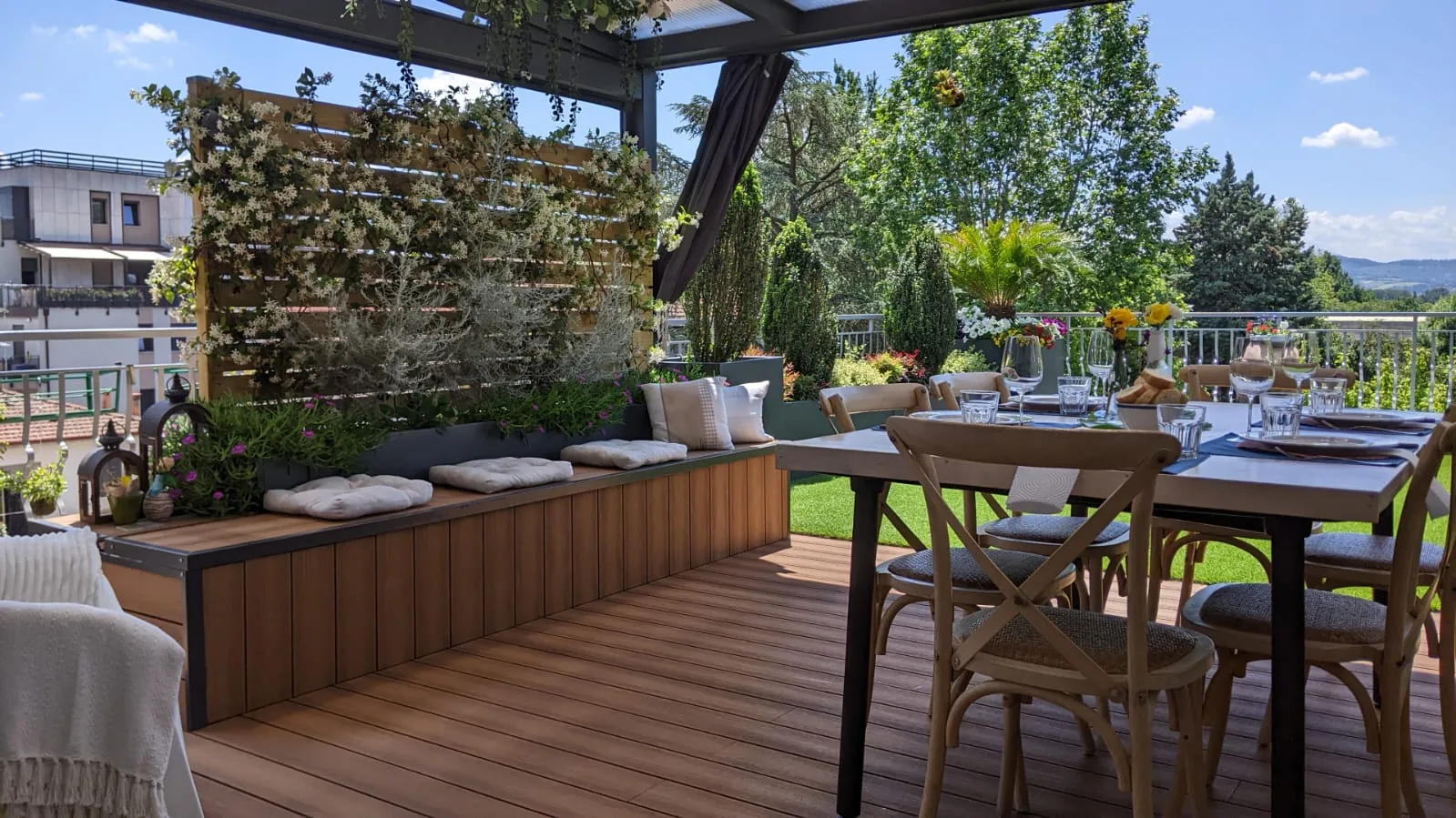 Tavolo apparecchiato sotto una pergola scura, con sedie in legno, una panca a cassone, cuscini e una parete di rampicanti su una terrazza