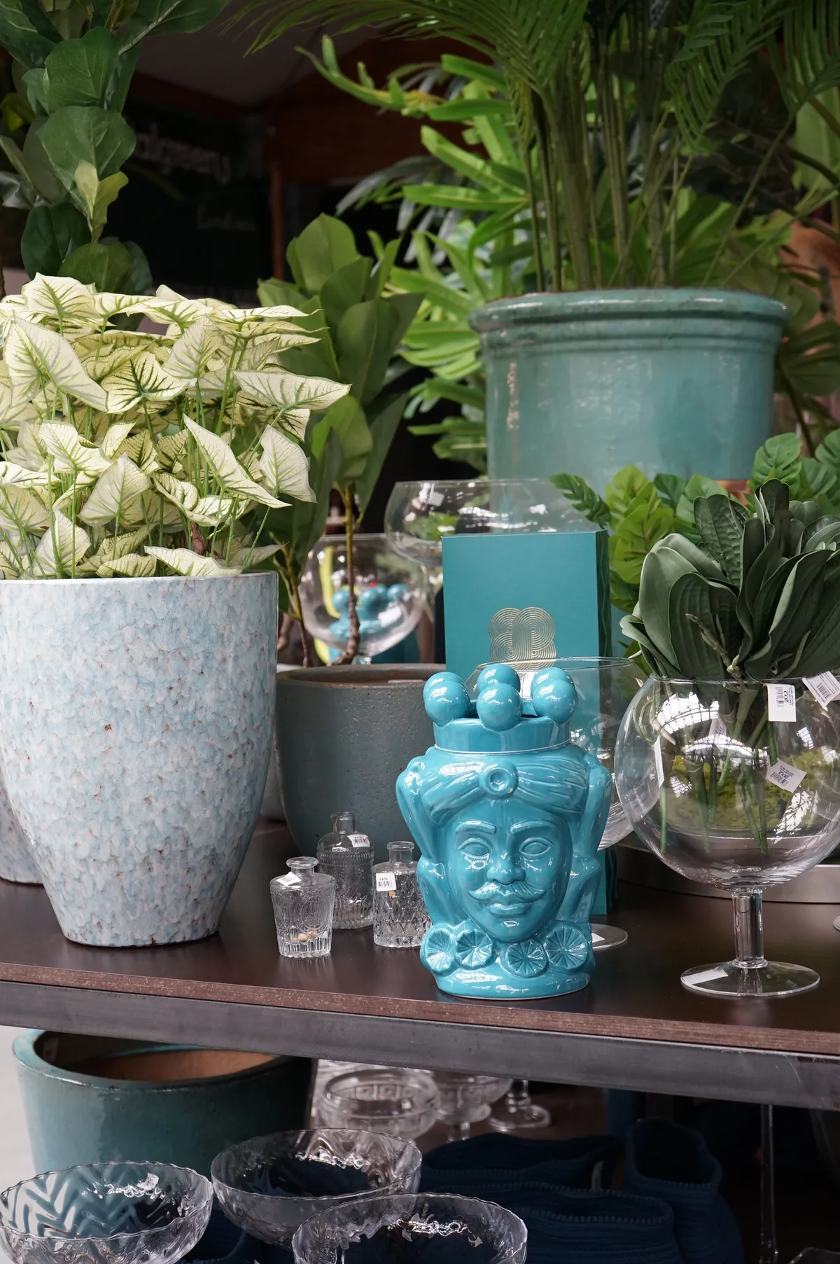 Portavasi in ceramica smaltata chiara e verde acqua con piante a foglia variegata, un vaso a testa azzurro e bicchieri di vetro