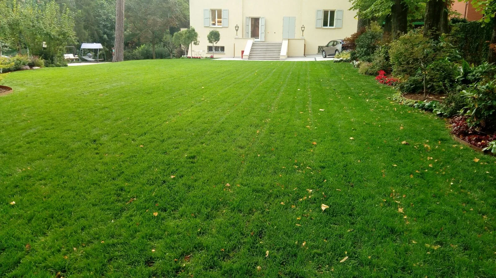 Grande prato tagliato a strisce davanti a una villa color crema, con aiuole fiorite e alberi lungo i lati