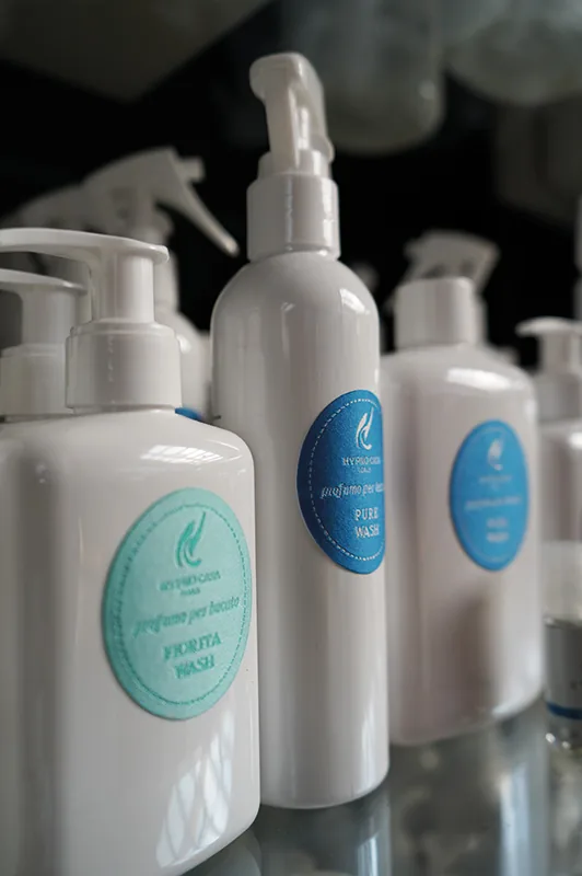 Flaconi bianchi di profumo per bucato con il dosatore a pompa ed etichette azzurre e blu, allineati su un ripiano di vetro, con altri flaconi spray dietro