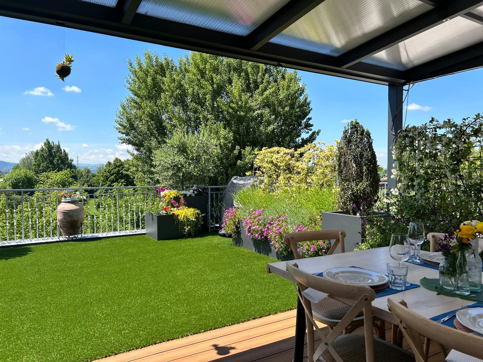Terrazza con prato sintetico, un tavolo apparecchiato sotto la pergola, fioriere fiorite e alberi sullo sfondo