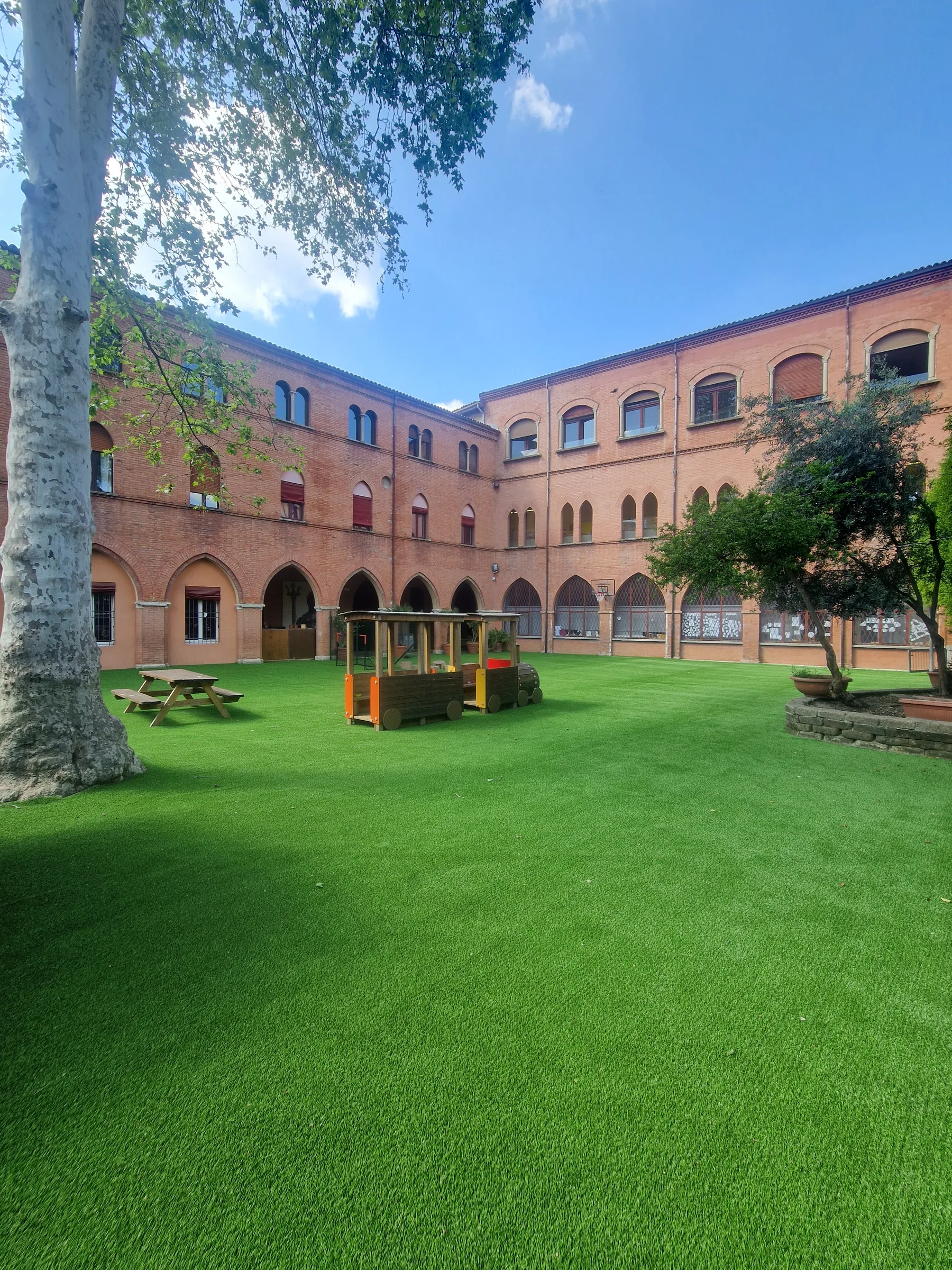Cortile di un edificio in mattoni con portico ad archi, coperto di prato sintetico, con un tavolo da pic-nic e un trenino di legno per giocare