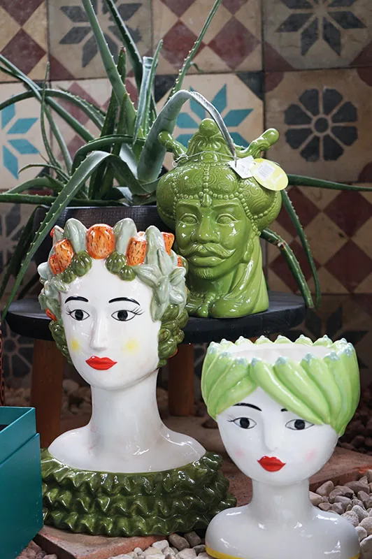 Due vasi a testa di donna e una testa d'uomo verde in ceramica colorata, con un'aloe alle spalle, davanti a un muro di cementine decorate