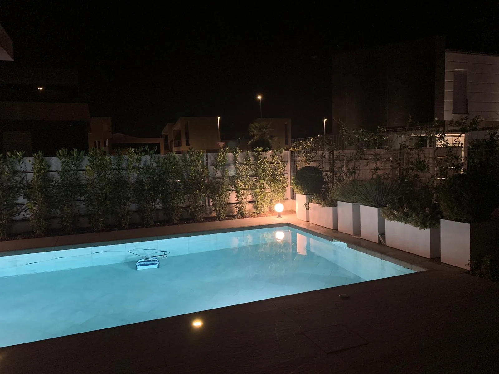 Piscina illuminata di notte, con fioriere bianche e una siepe lungo il bordo