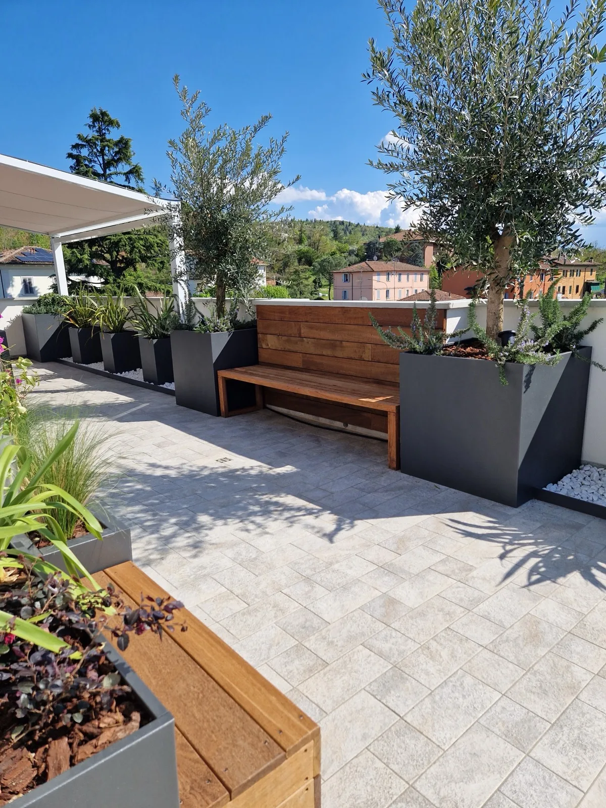 Terrazza con fioriere antracite, una panca in legno con lo schienale fra le fioriere e due olivi con il rosmarino alla base, con le colline sullo sfondo