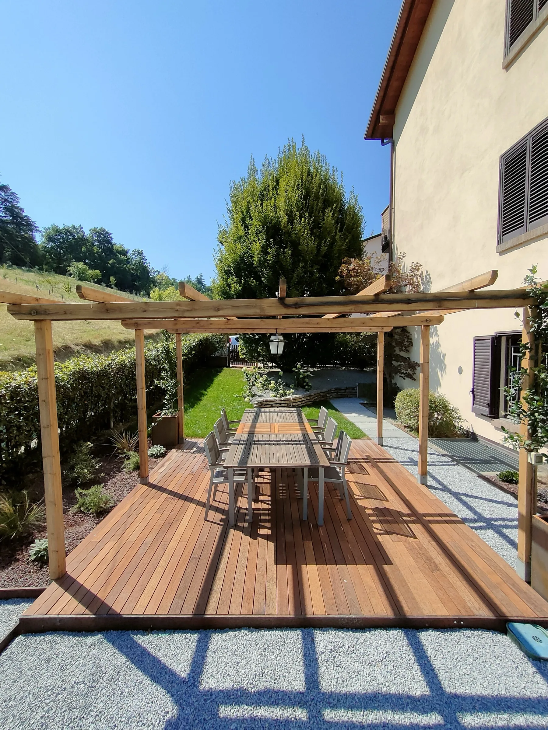 Pergolato in legno chiaro su una pedana, con un tavolo lungo, accanto a una casa in campagna