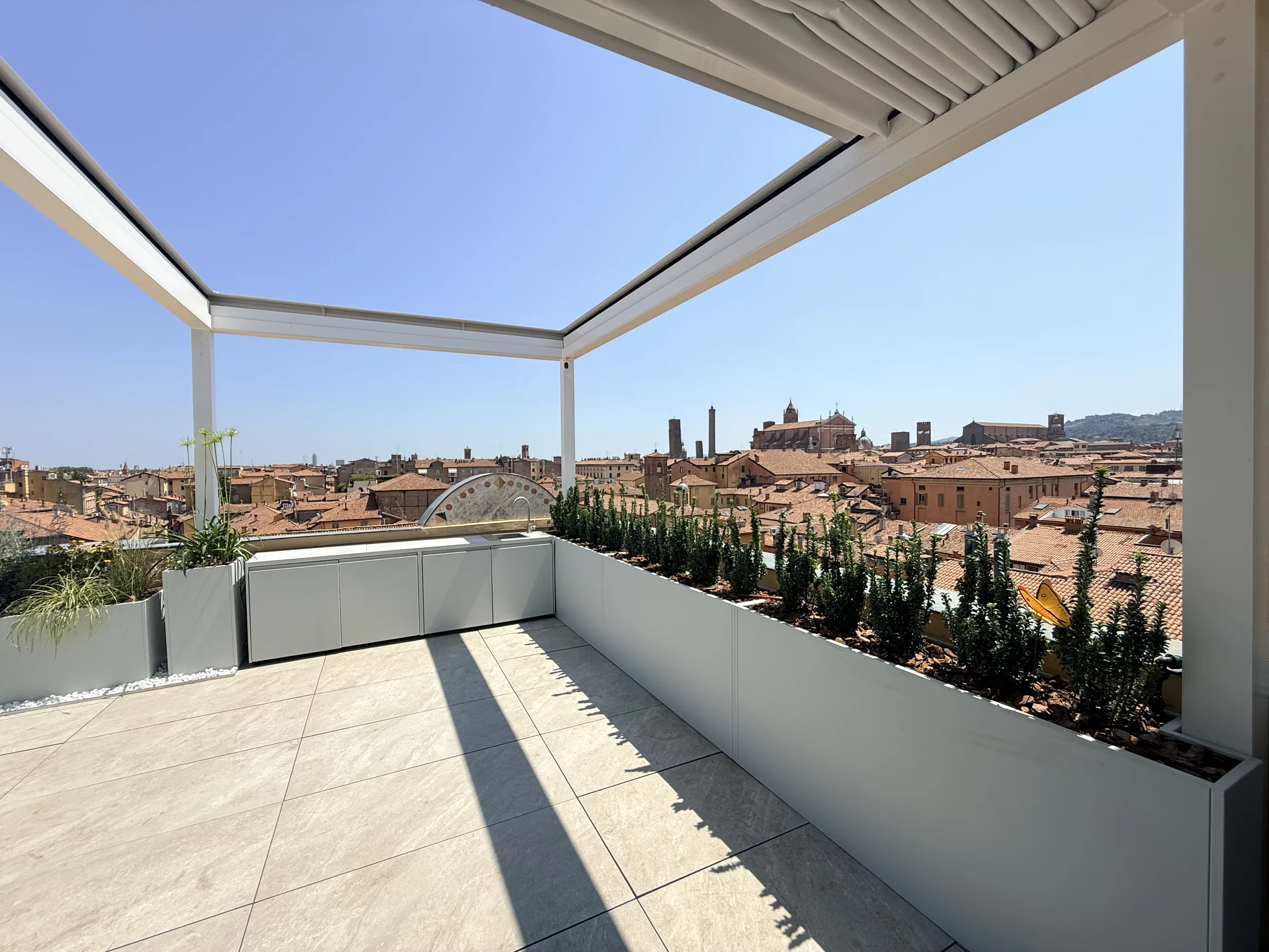 Terrazza all'ultimo piano con pergola bioclimatica a lamelle, fioriere continue di arbusti colonnari e i tetti della città con le due torri sul fondo