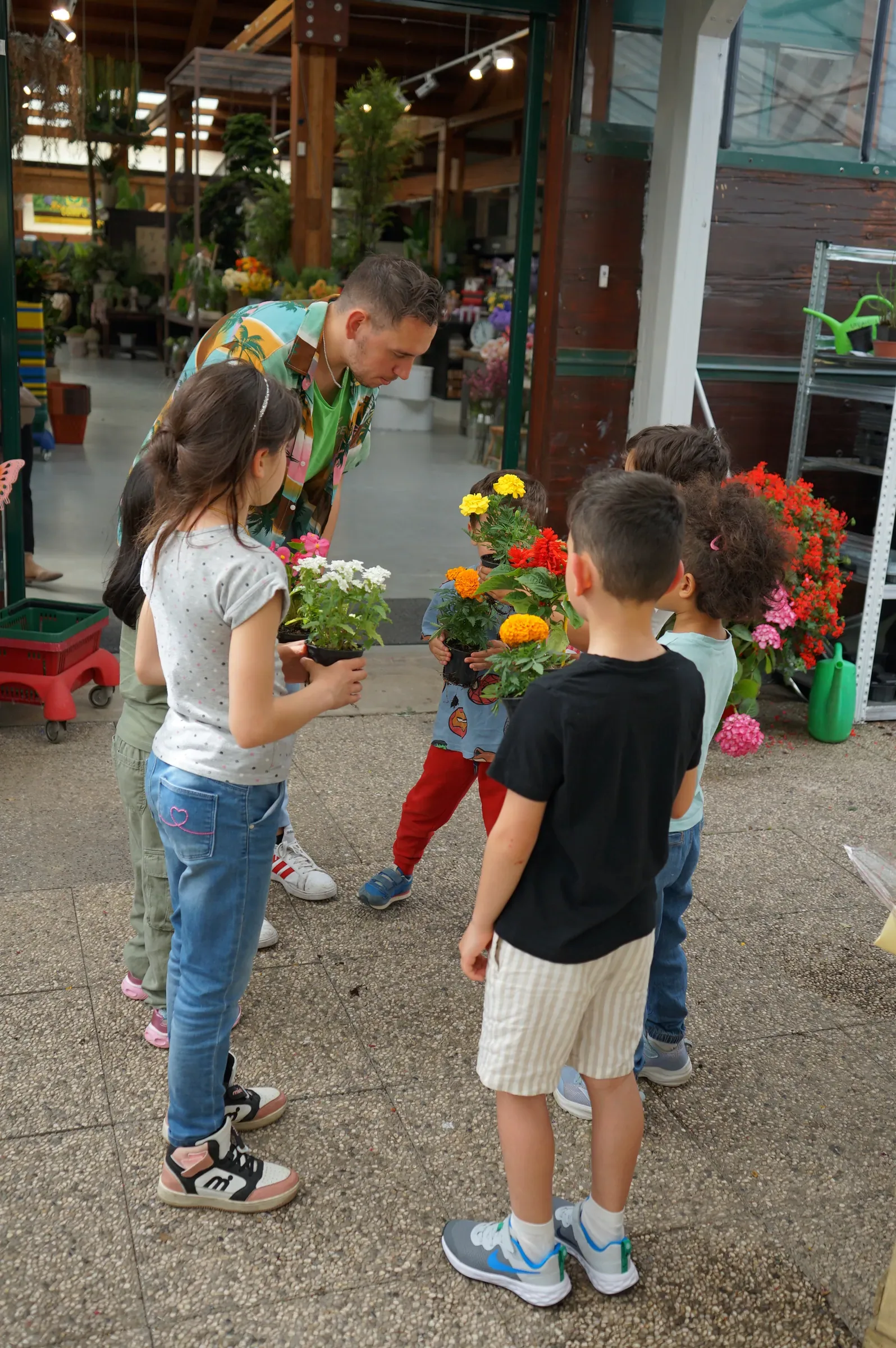 Bambini con i vasetti di fiori in mano fra i banchi del garden, con un ragazzo in camicia colorata chinato verso di loro