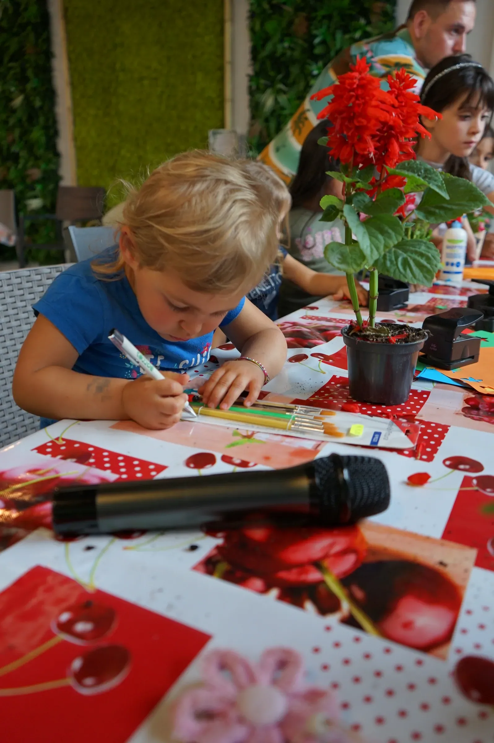 Un bimbo colora un cartoncino con il pennarello, al tavolo con la tovaglia a ciliegie, accanto a una salvia rossa in vaso