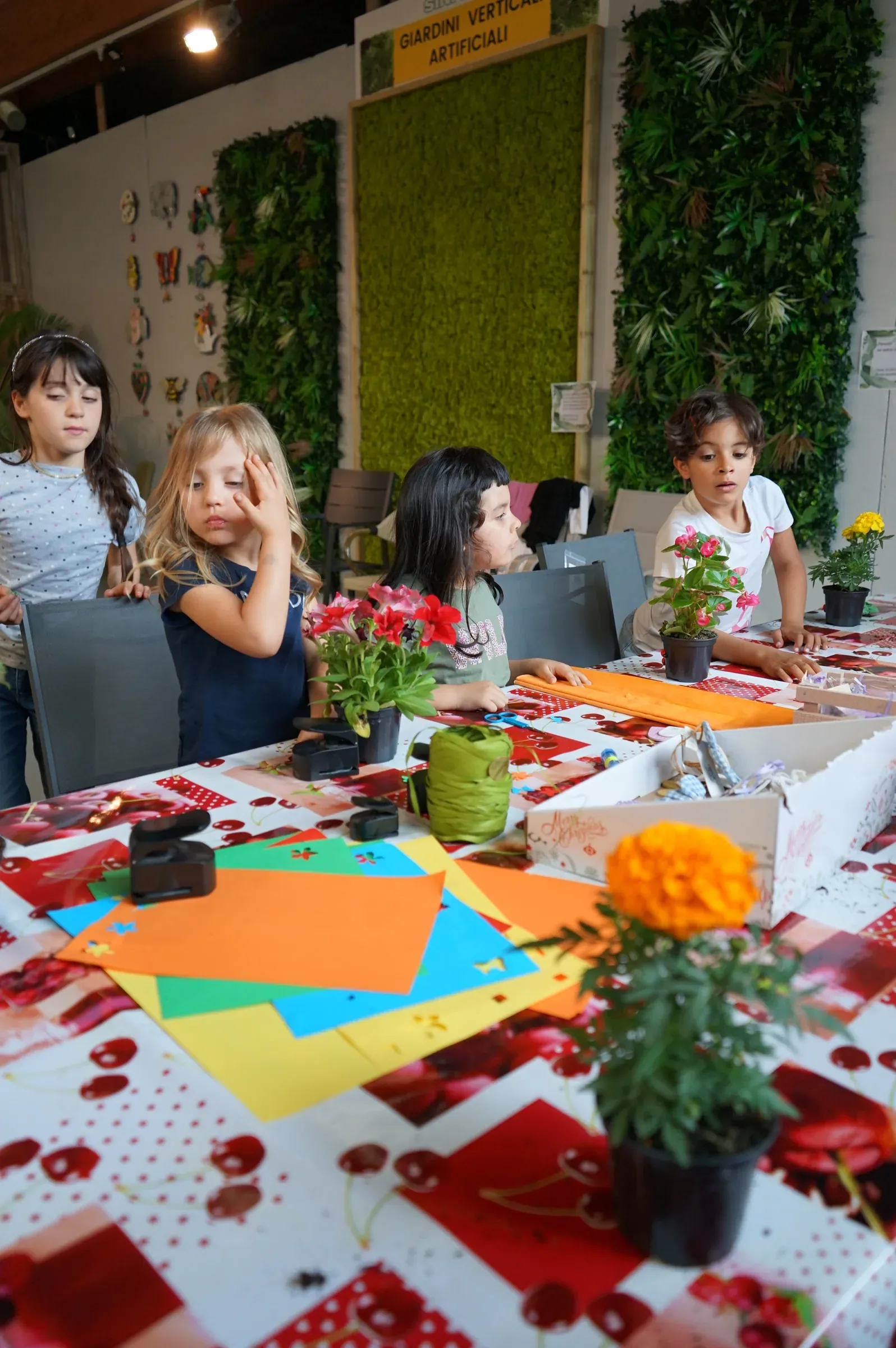 Bambini al tavolo fra cartoncini colorati e vasetti di fiori, davanti alle pareti di verde
