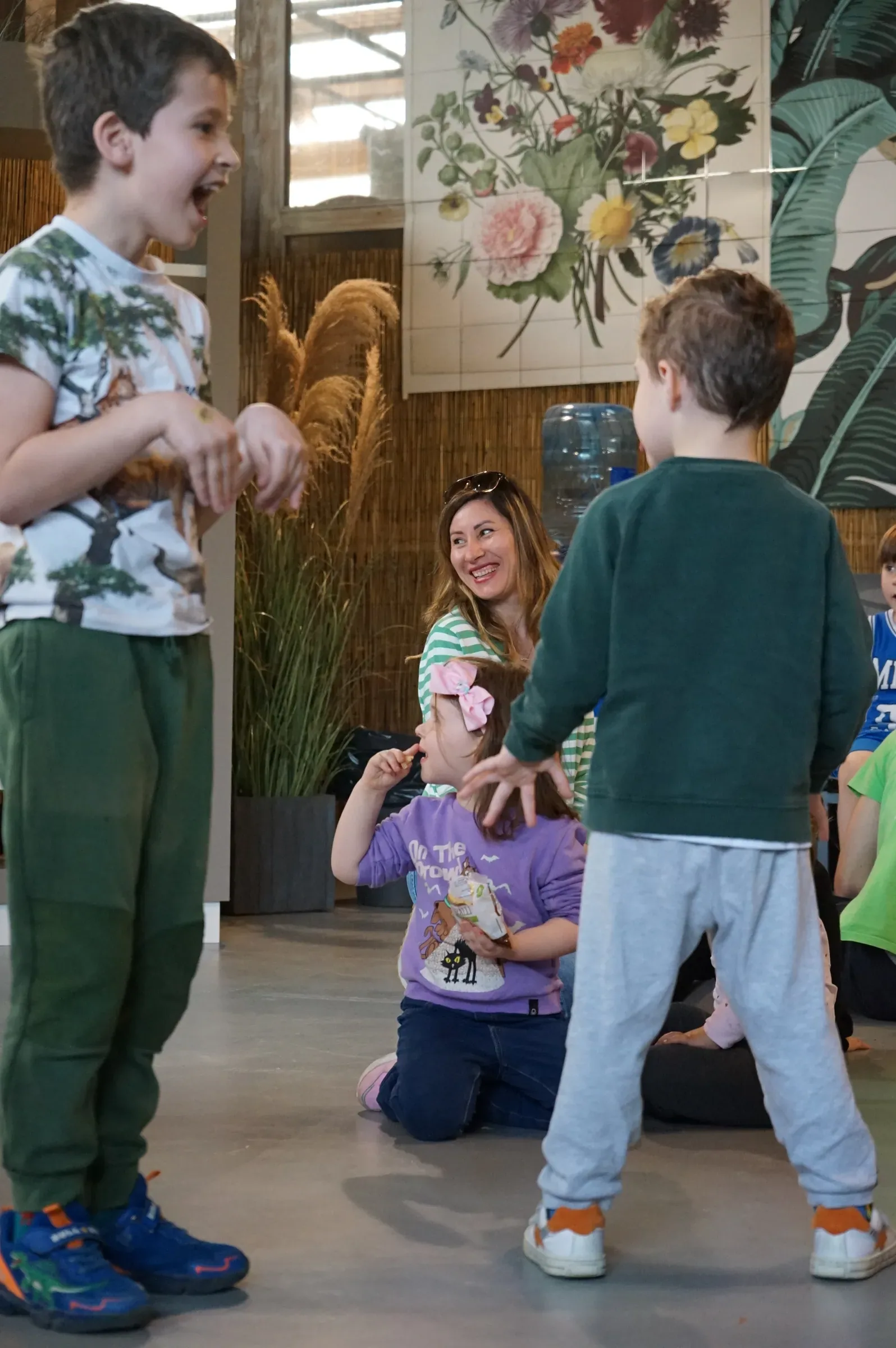 Un bambino con la maglietta stampata ad alberi ride a bocca aperta; accanto a lui un bambino di spalle e una ragazza che sorride a una bambina seduta a terra