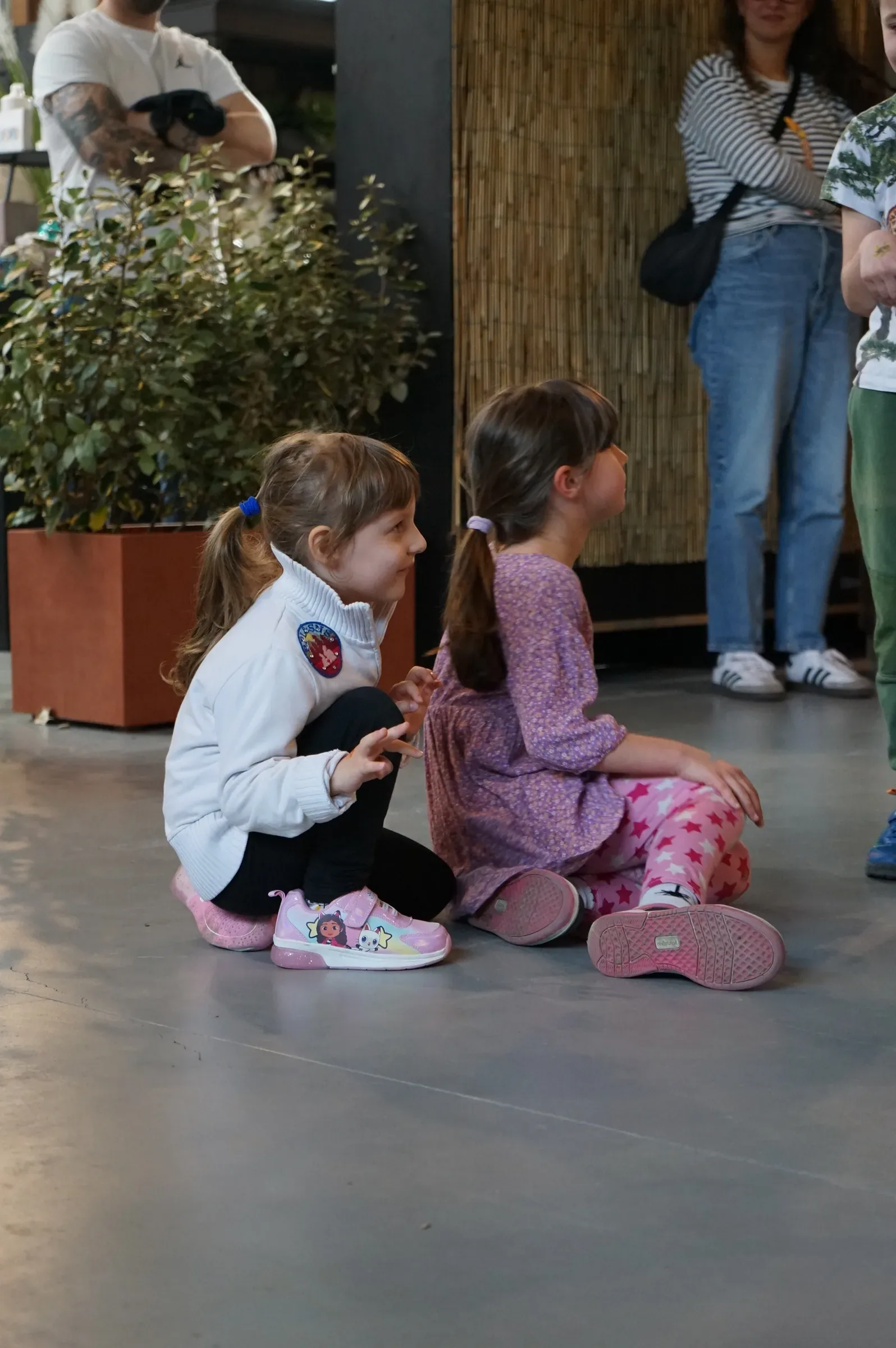 Due bambine sedute sul pavimento grigio, una sorride girata verso l'altra che guarda altrove, accanto a un grande vaso con una pianta