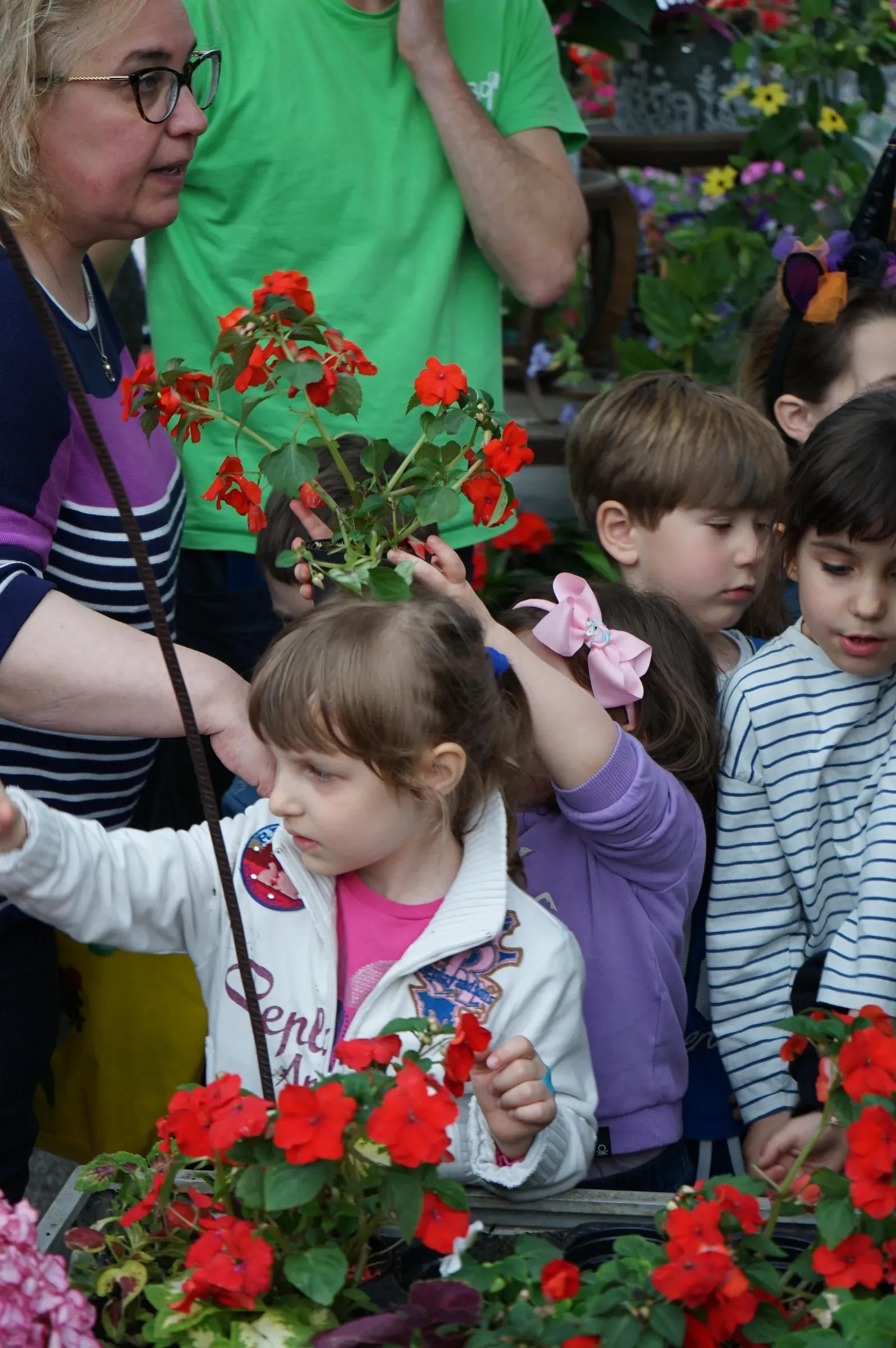 Una bambina con la felpa bianca fra altri bambini davanti a un banco di fiori rossi; una compagna alza una piantina fiorita, accanto a una signora con gli occhiali