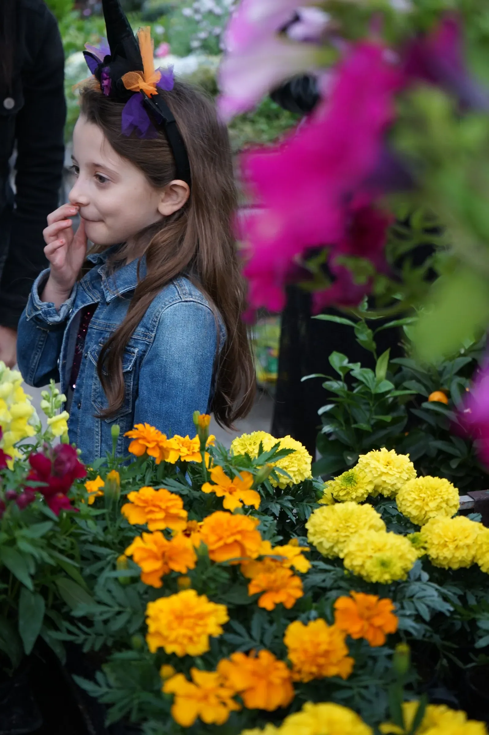 Una bambina con il cerchietto da strega sorride con la mano vicino alla bocca, dietro un banco di tageti gialli e arancioni