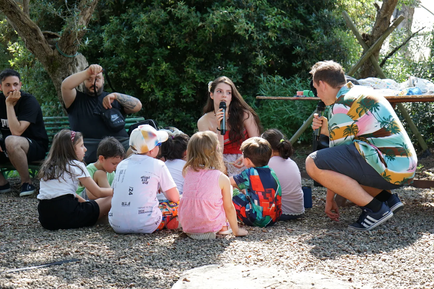 Vaiana e un animatore accovacciato, tutti e due con il microfono, davanti ai bambini seduti sulla ghiaia all'ombra degli alberi